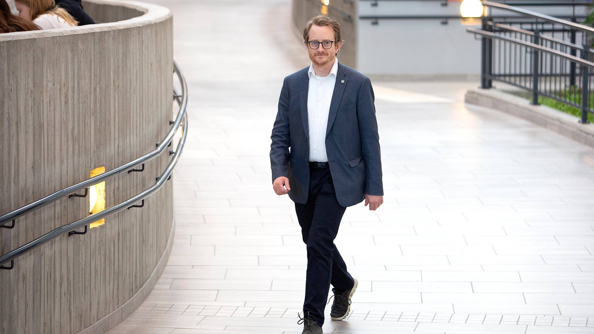 En mann i dress går forbi en buet betongvegg i en offentlig bygning. I bakgrunnen sitter flere personer på en benk. Gulvflisene er lyse, og det er lamper langs veggen. Omgivelsene tyder på et moderne og urbant miljø. (Bildebeskrivelsen er laget av en KI-tjeneste)