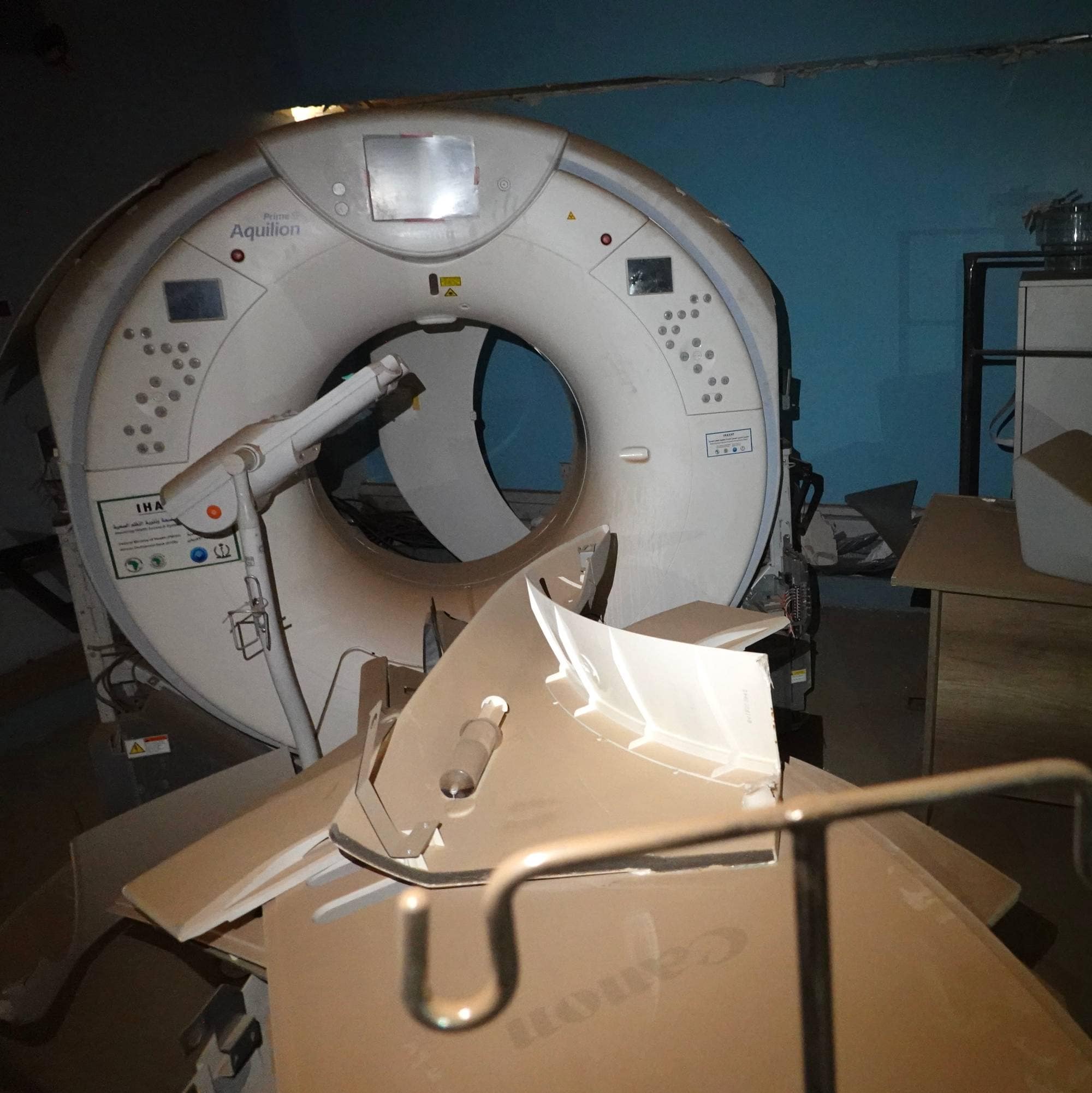 Et CT-apparat står i et rom med blåmalte vegger. Det ligger bygningsrester foran apparatet. Til høyre for apparatet står et hvitt skap på et stativ. Det ligger også noe på skapet. (Bildebeskrivelsen er laget av en KI-tjeneste)