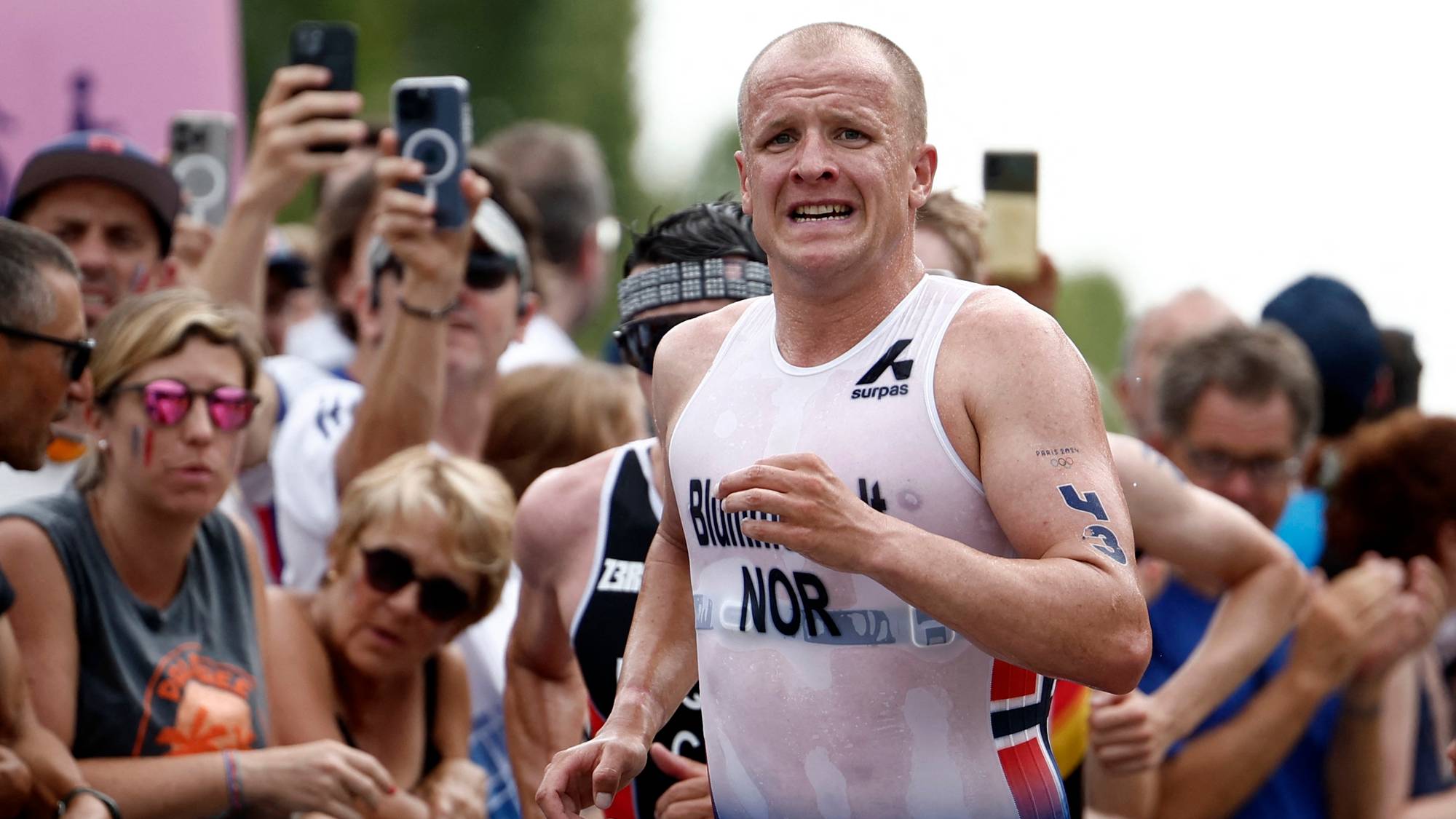 En mann i triatlonsdrakt løper foran et publikum. Han har en svett overkropp og ser utmattet ut. Bak ham står folk med telefoner oppe. (Bildebeskrivelsen er laget av en KI-tjeneste)