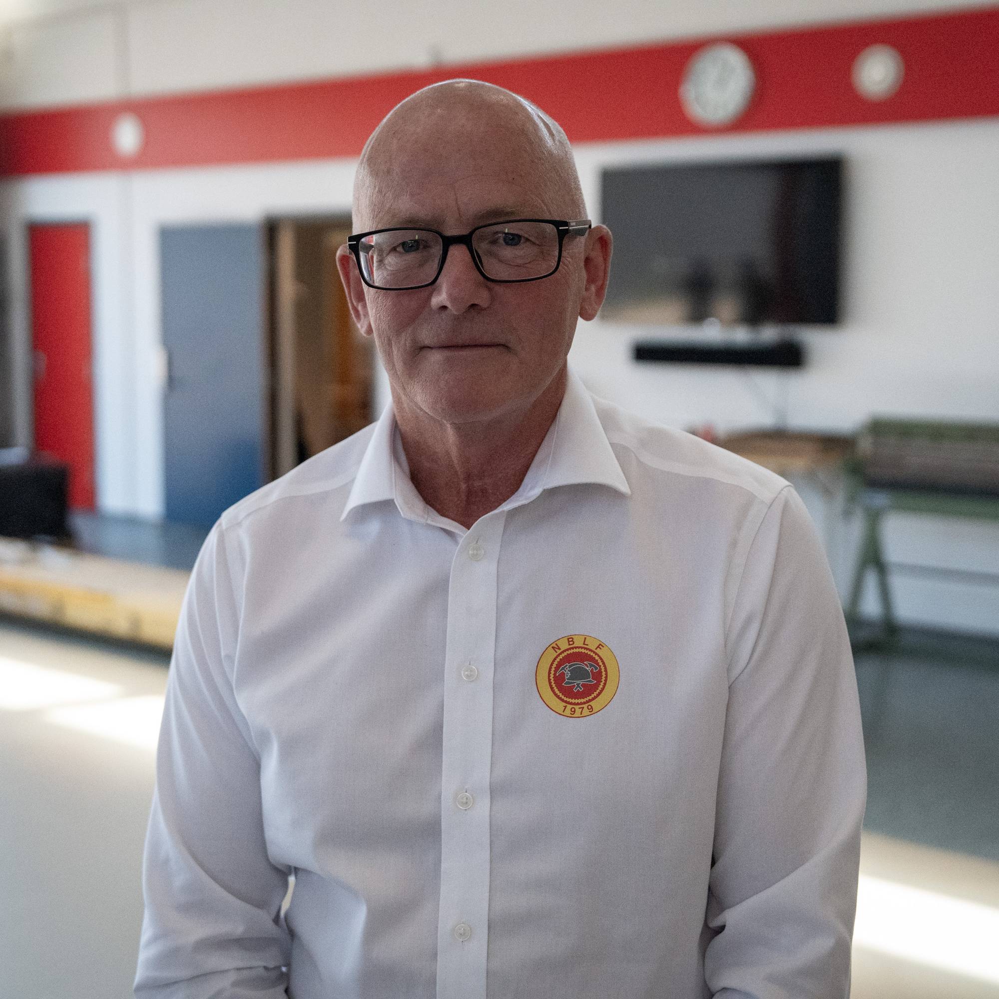 Mann med briller og hvit skjorte med NBLF-logo står inne i en brannstasjon. (Bildebeskrivelsen er laget av en KI-tjeneste)