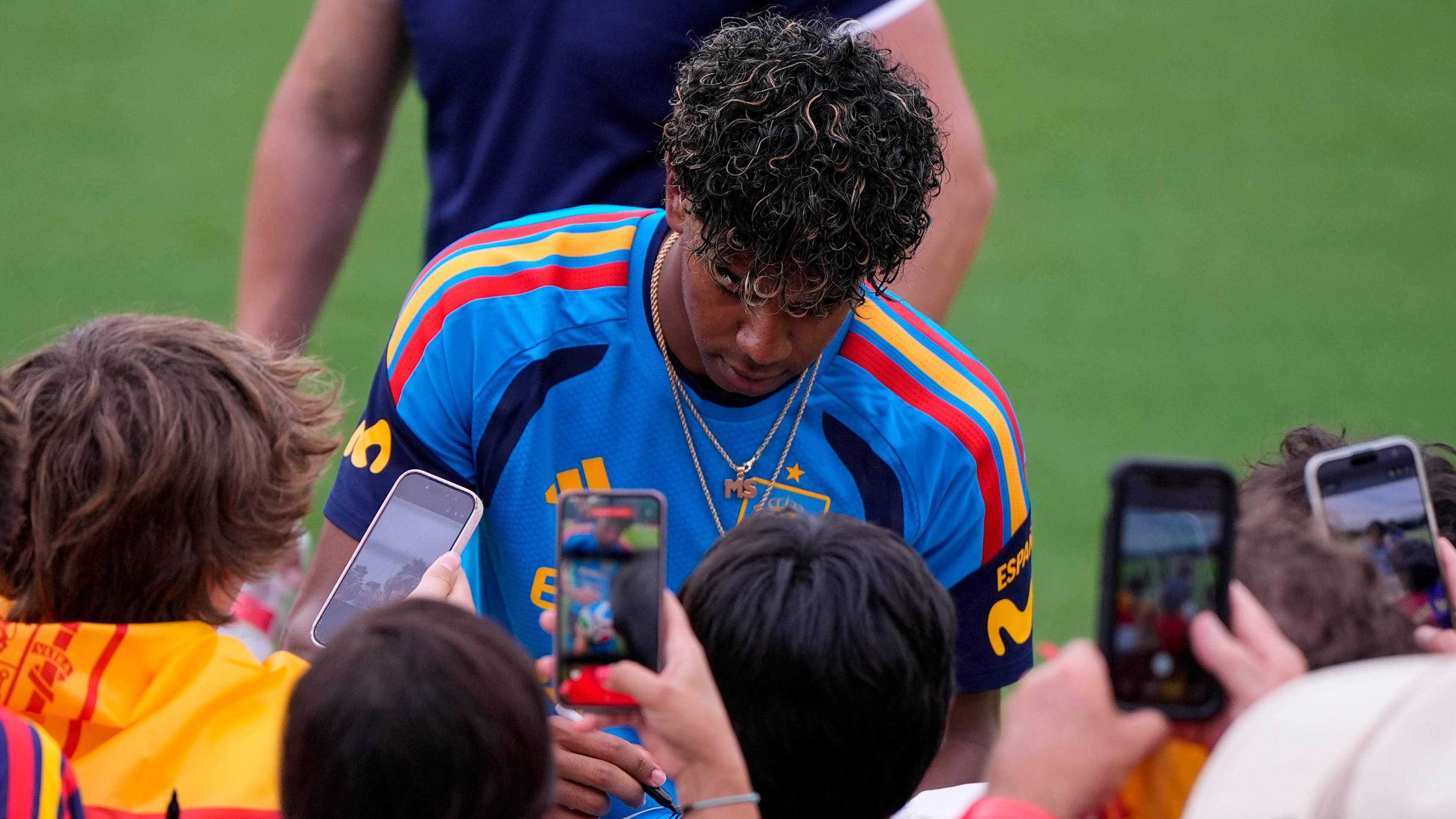 En fotballspiller i blå drakt med gule og røde striper signerer autografer. Rundt spilleren står flere personer som holder opp telefoner for å ta bilder. Spilleren har på seg et smykke med bokstavene "MS". (Bildebeskrivelsen er laget av en KI-tjeneste)