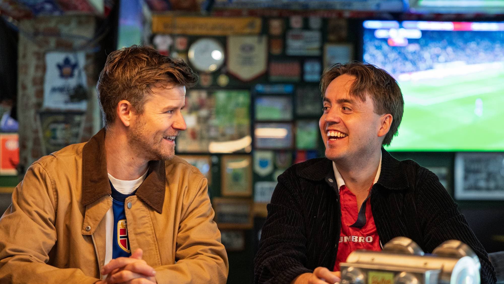 Nicolay Ramm og Markus Neby sitter ved et bord på en pub og ler. Den ene mannen har på seg en lys jakke og en blå trøye, mens den andre har på seg en mørk jakke over en rød trøye. Bak dem er det dekorasjoner og en TV som viser en fotballkamp. 