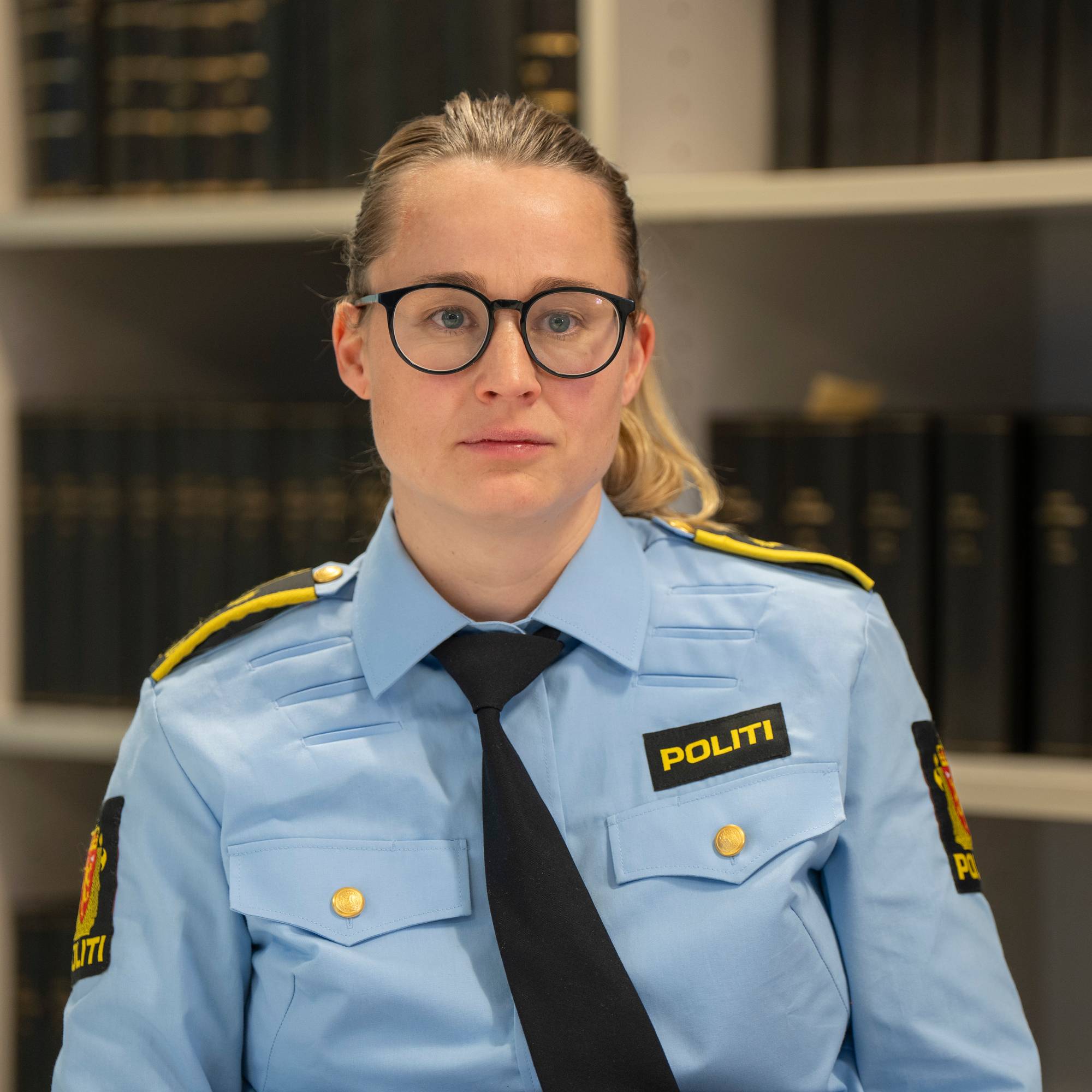 En kvinne med briller og en politi-skjorte sitter foran en bokhylle full av svarte bøker.