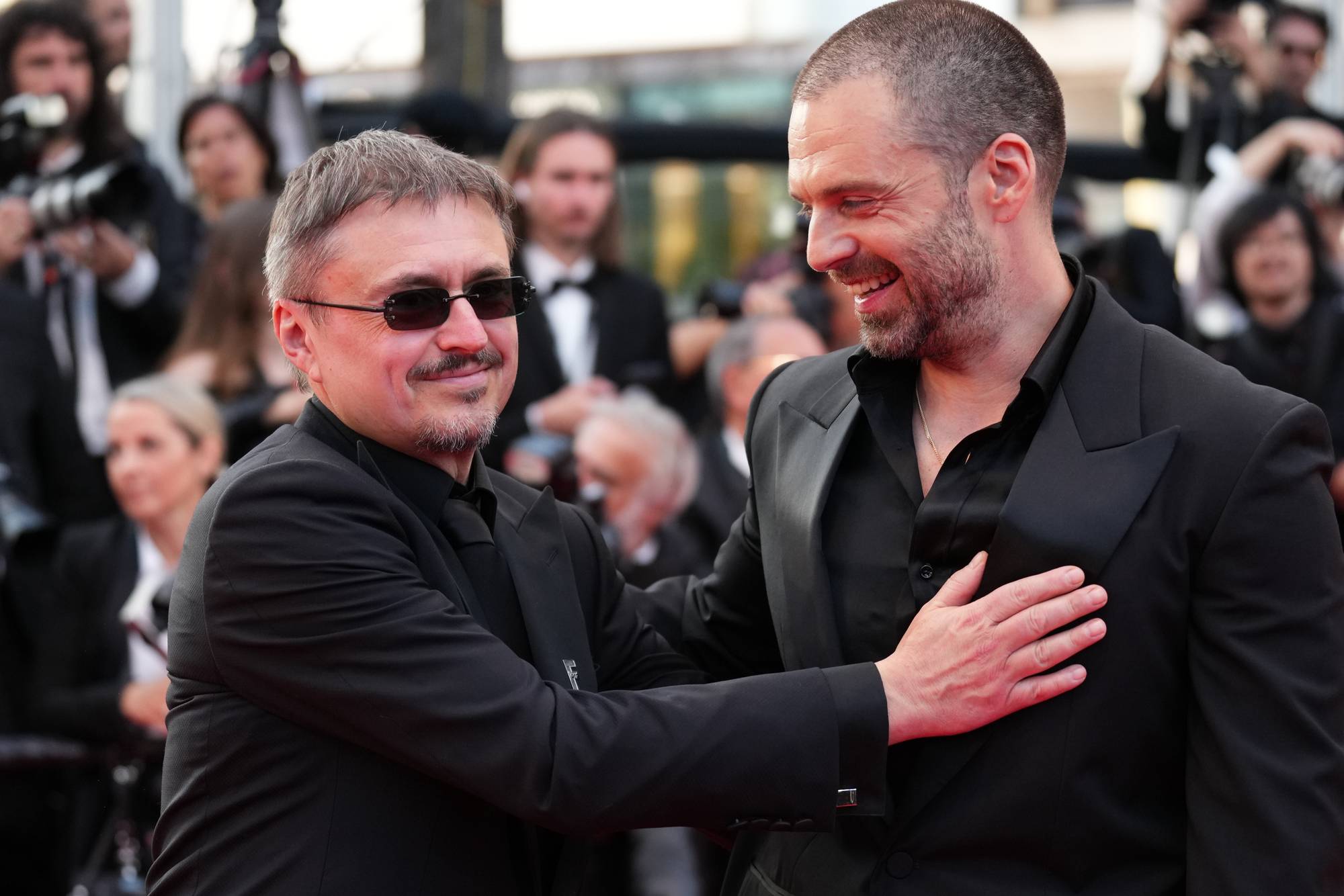 To menn i dress smiler og snakker med hverandre på en rød løper. Den ene mannen, med solbriller, legger hånden på brystet til den andre. De er omringet av andre mennesker og fotografer i bakgrunnen. (Bildebeskrivelsen er laget av en KI-tjeneste)