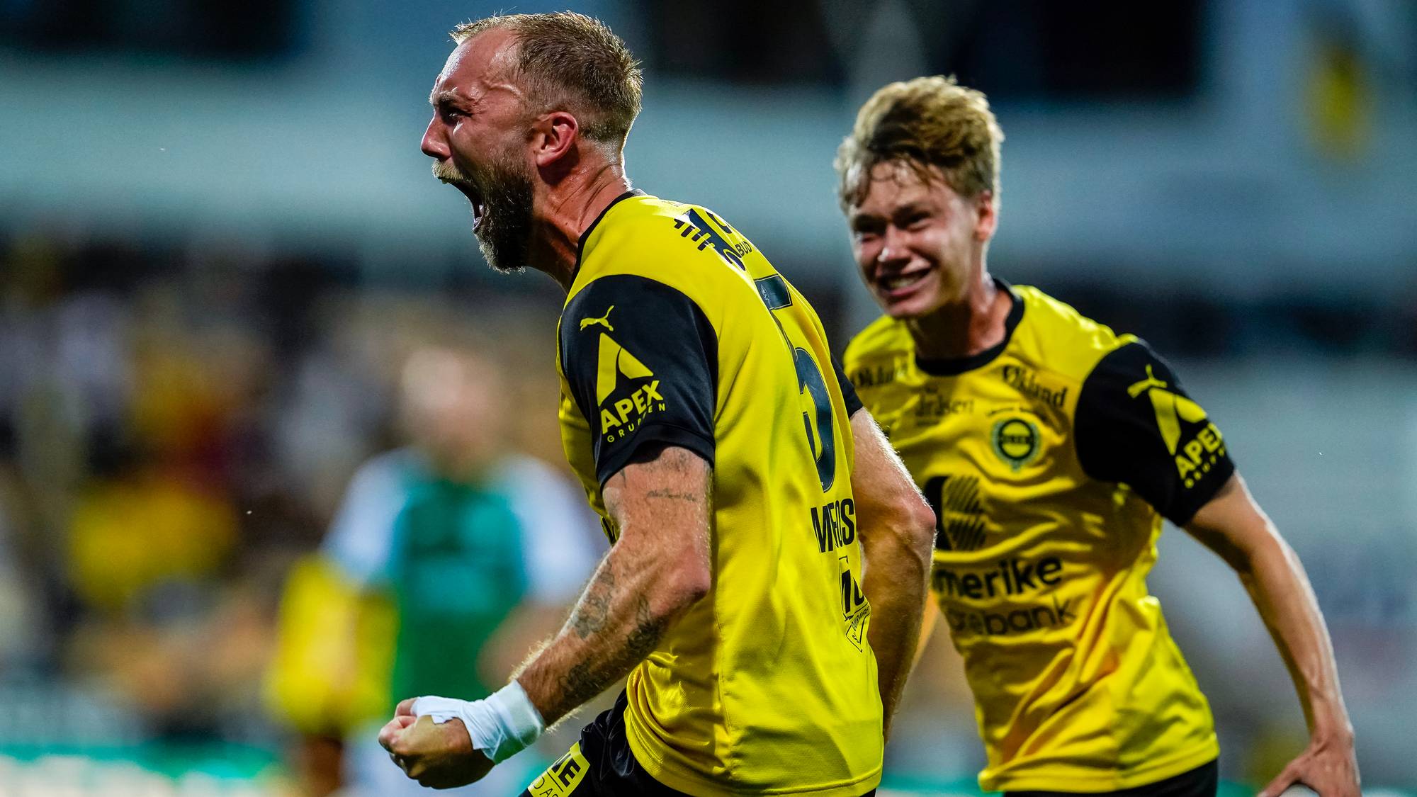Fotballspiller jubler med knyttet neve etter mål, etterfulgt av en lagkamerat. (Bildebeskrivelsen er laget av en KI-tjeneste)