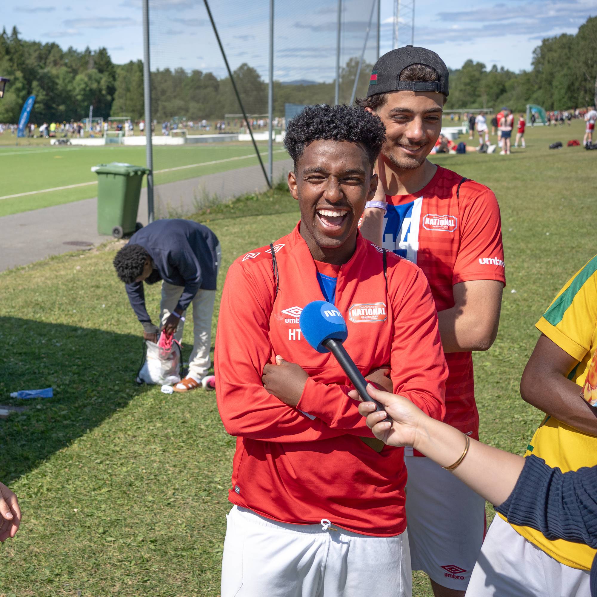 Tre fotballspillere i røde drakter intervjues på en fotballbane utendørs. (Bildebeskrivelsen er laget av en KI-tjeneste)