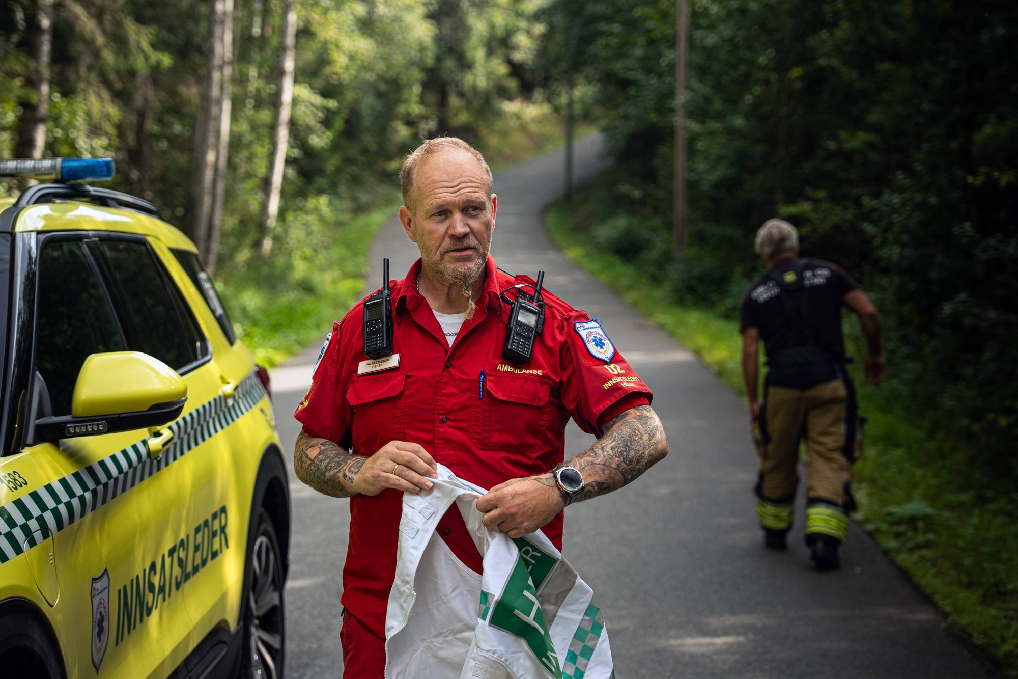 En ambulanse-innsatsleder ved en gul bil på en skogsvei, med en brannkonstabel i bakgrunnen. (Bildebeskrivelsen er laget av en KI-tjeneste)