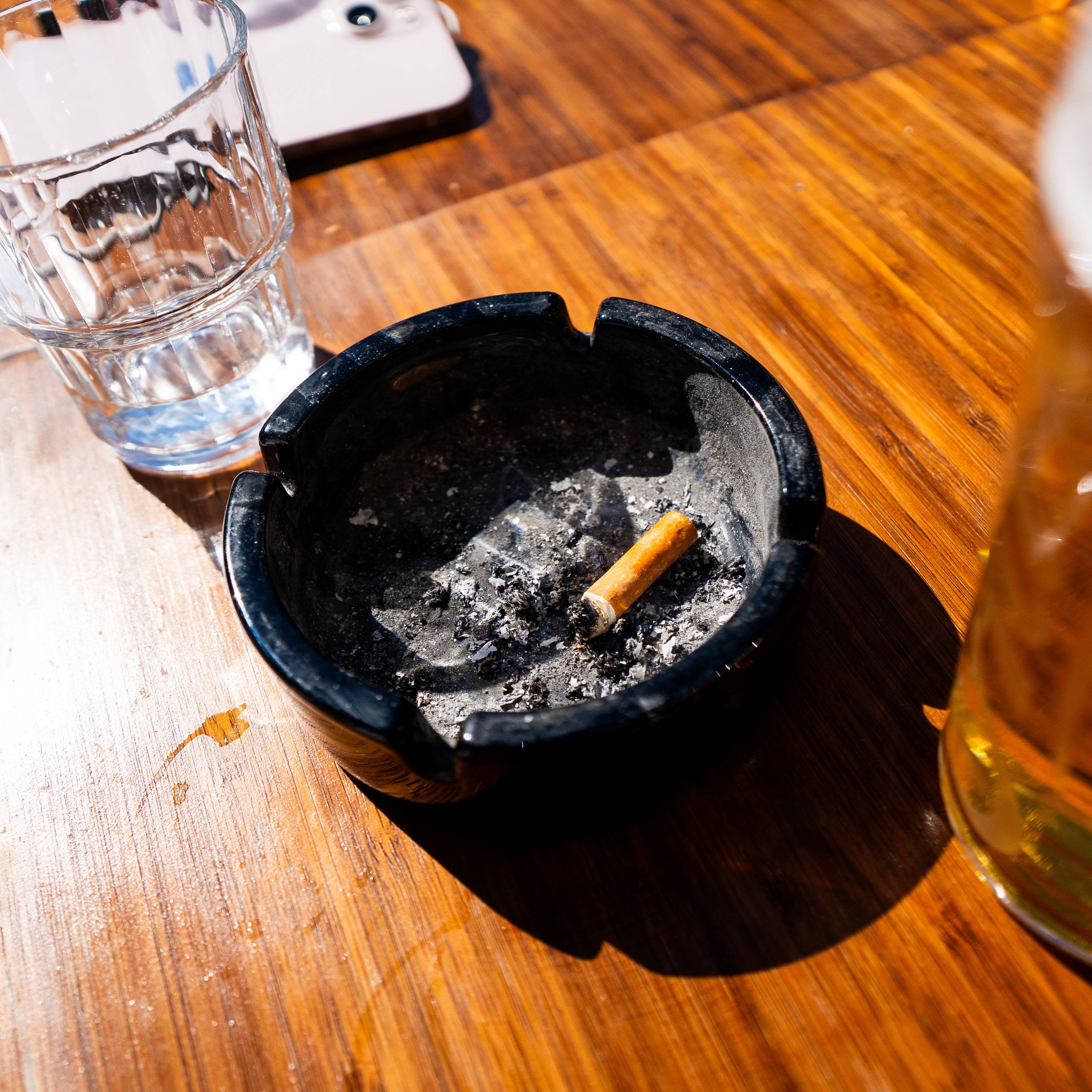 Et askfat med et sigarettstump ligg på et trebord. Ved sidan av askfatet står et glas med vann og et ølglas. Ein lys skjerm av ein mobiltelefon ligg i bakgrunnen. (Bildebeskrivelsen er laget av ein KI-teneste)