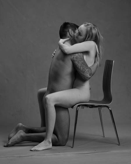 En naken dame med lyst hår sitter på en stol og holder rundt en naken mann med mørkt hår og tatoveringer som står på kne mellom bena hennes