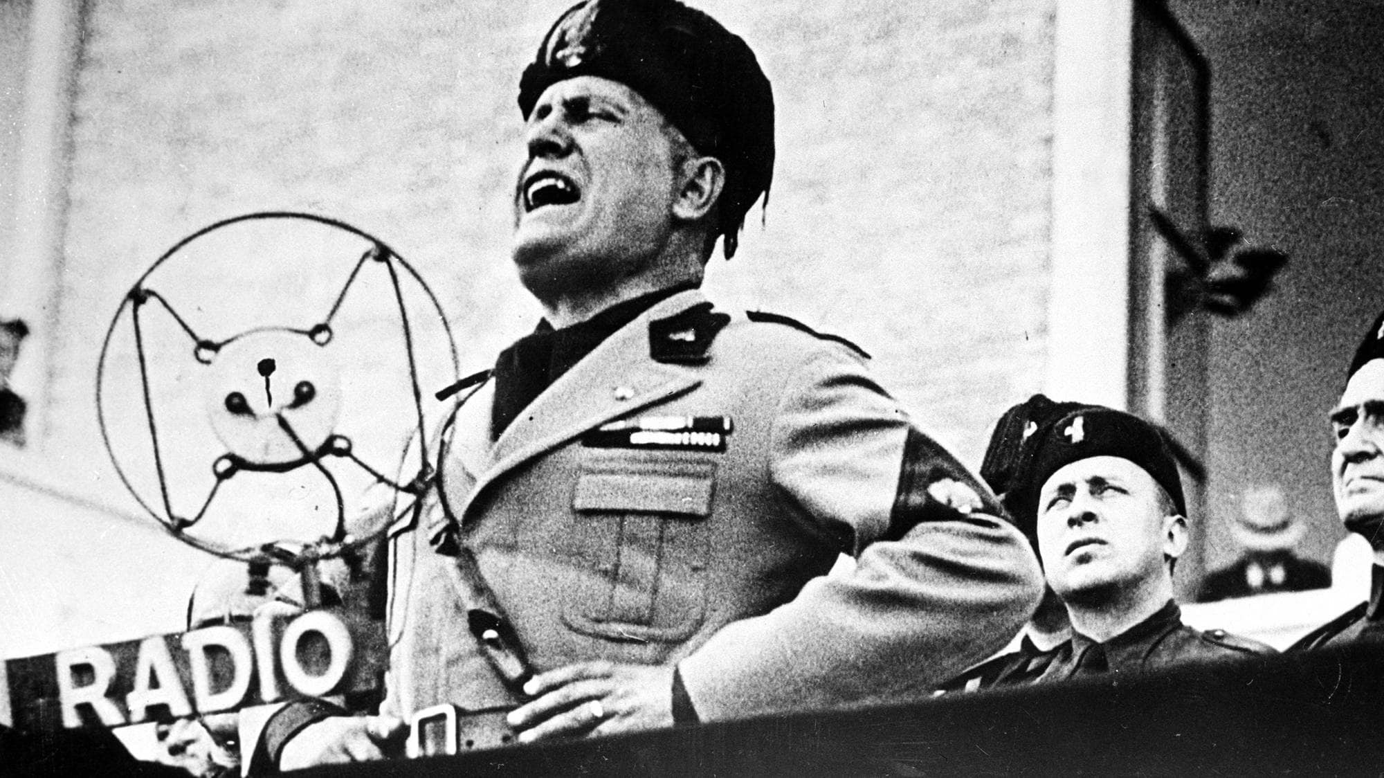 Mann i uniform taler ved en mikrofon mens flere menn i uniform ser opp. En banner med teksten "GERMAN RADIO" er synlig. Bildet er svart-hvitt. (Bildebeskrivelsen er laget av en KI-tjeneste)