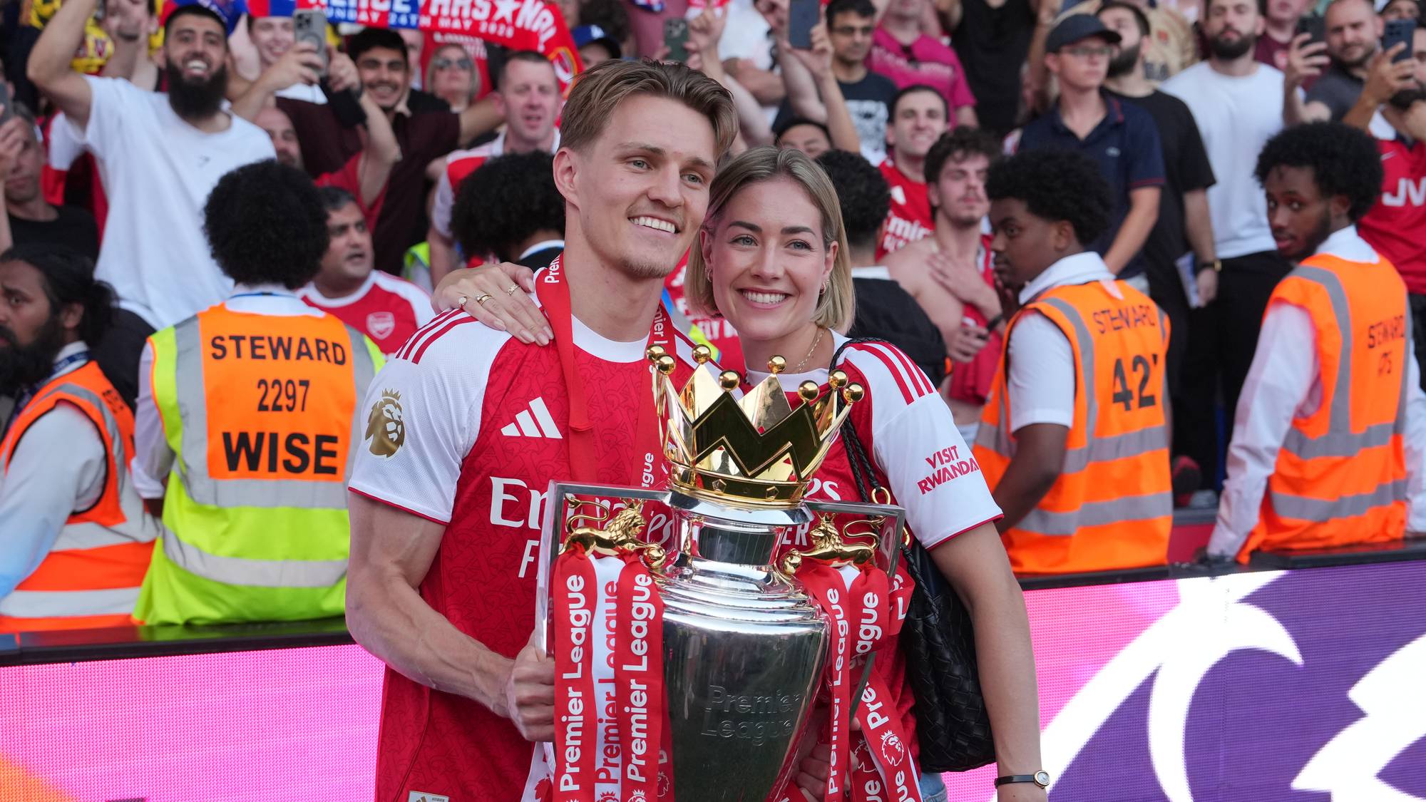 Martin Ødegaard og partneren holder Premier League-trofeet foran jublende publikum på en fotballstadion. (Bildebeskrivelsen er laget av en KI-tjeneste)