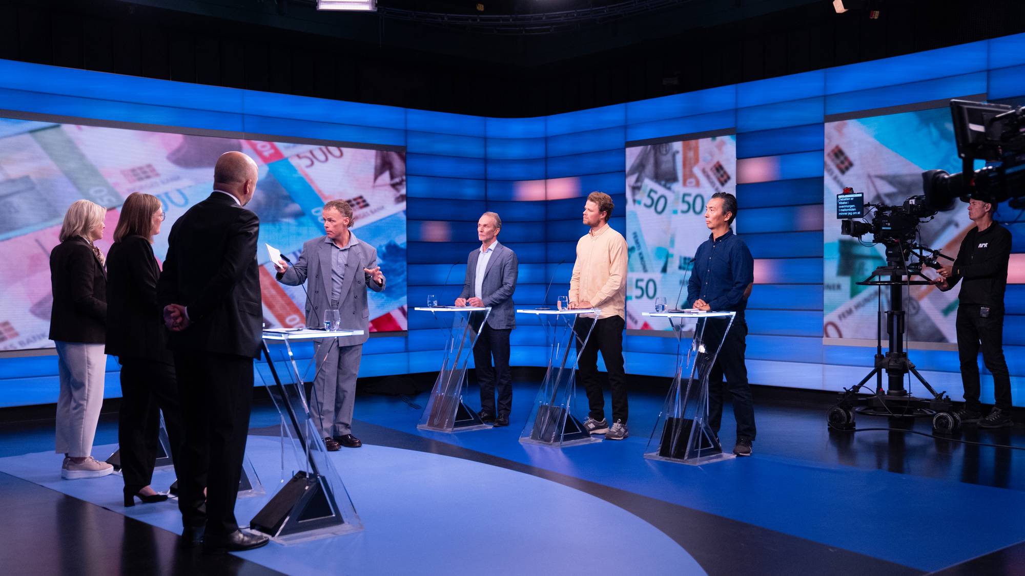 Debattdeltakere står ved talerstoler i et TV-studio med pengesedler på skjermene i bakgrunnen. (Bildebeskrivelsen er laget av en KI-tjeneste)