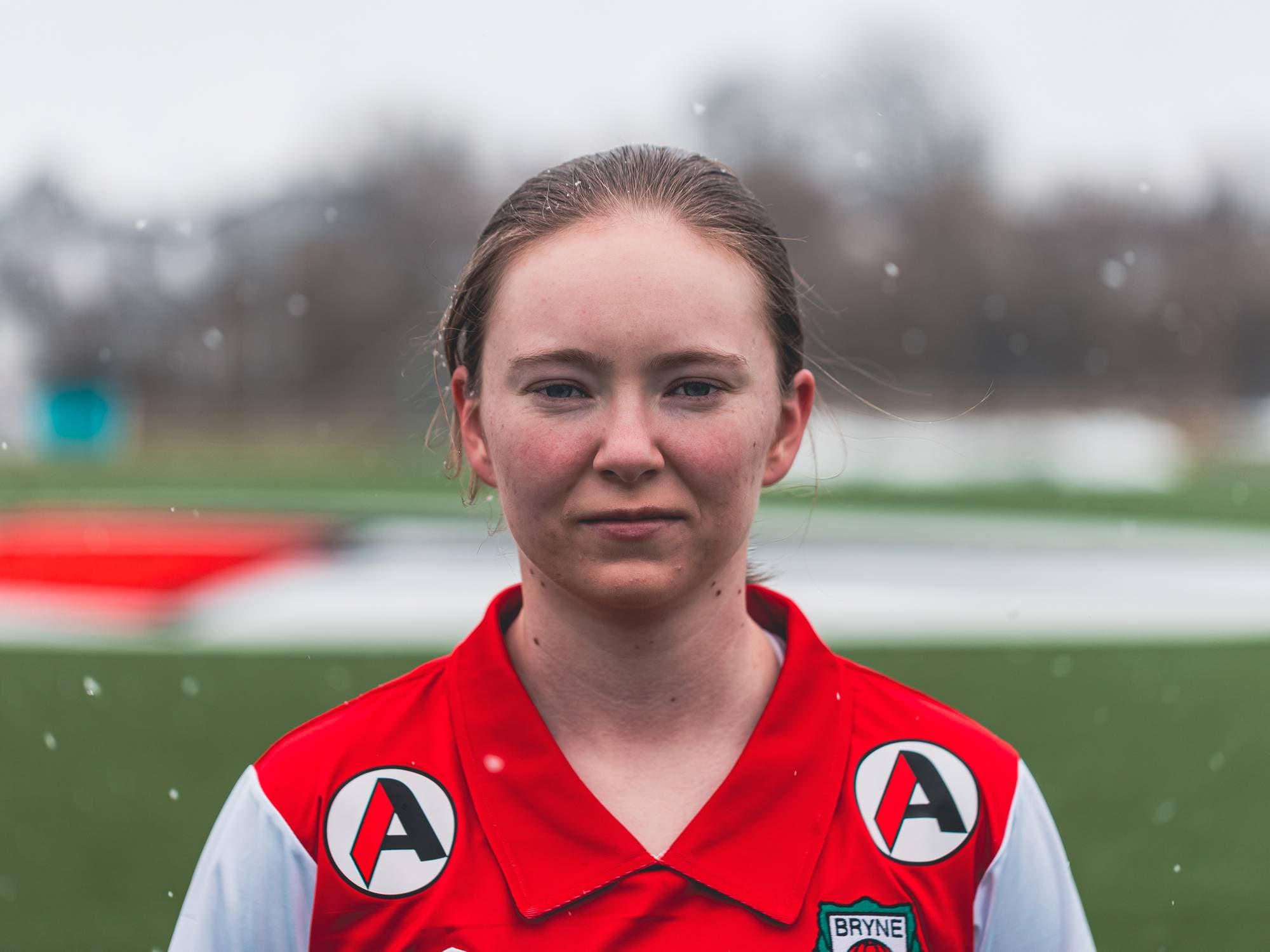 Et portrett av Karina. Hun ser alvorlig inn i kamera med sine lyse øyne, røde kinn og knallrøde Bryne FK fotballdrakt. Hun har rødlig blondt hår og vi ser snøen dale rundt henne ned på den grønne fotballbanen hun står på.