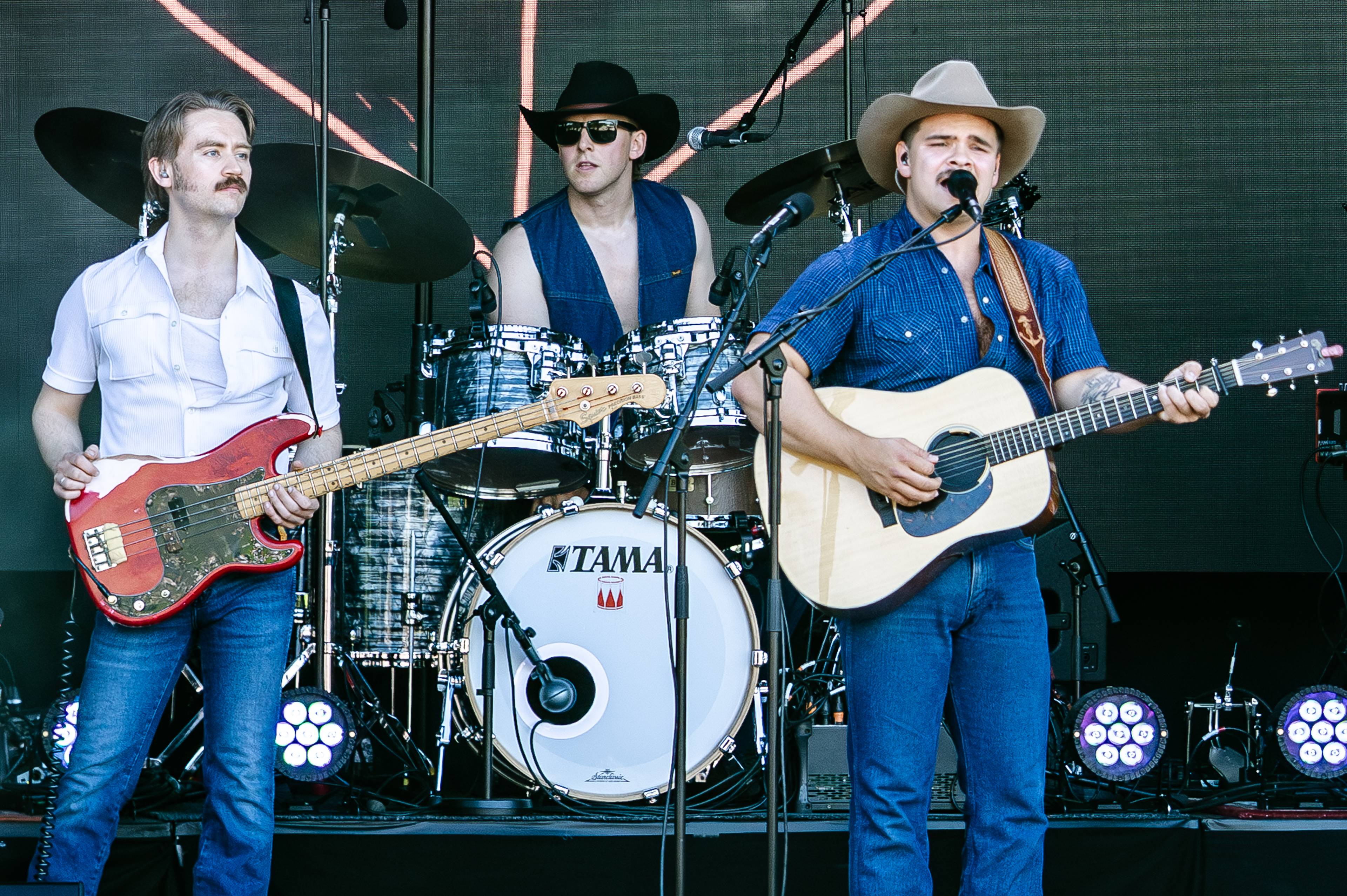 Tobias Sten med cowboyhatt spiller gitar mens han synger. En annen mann spiller bassgitar og en tredje sitter bak et trommesett. Musikere i cowboyantrekk opptrer på en scene. (Bildebeskrivelsen er laget av en KI-tjeneste)