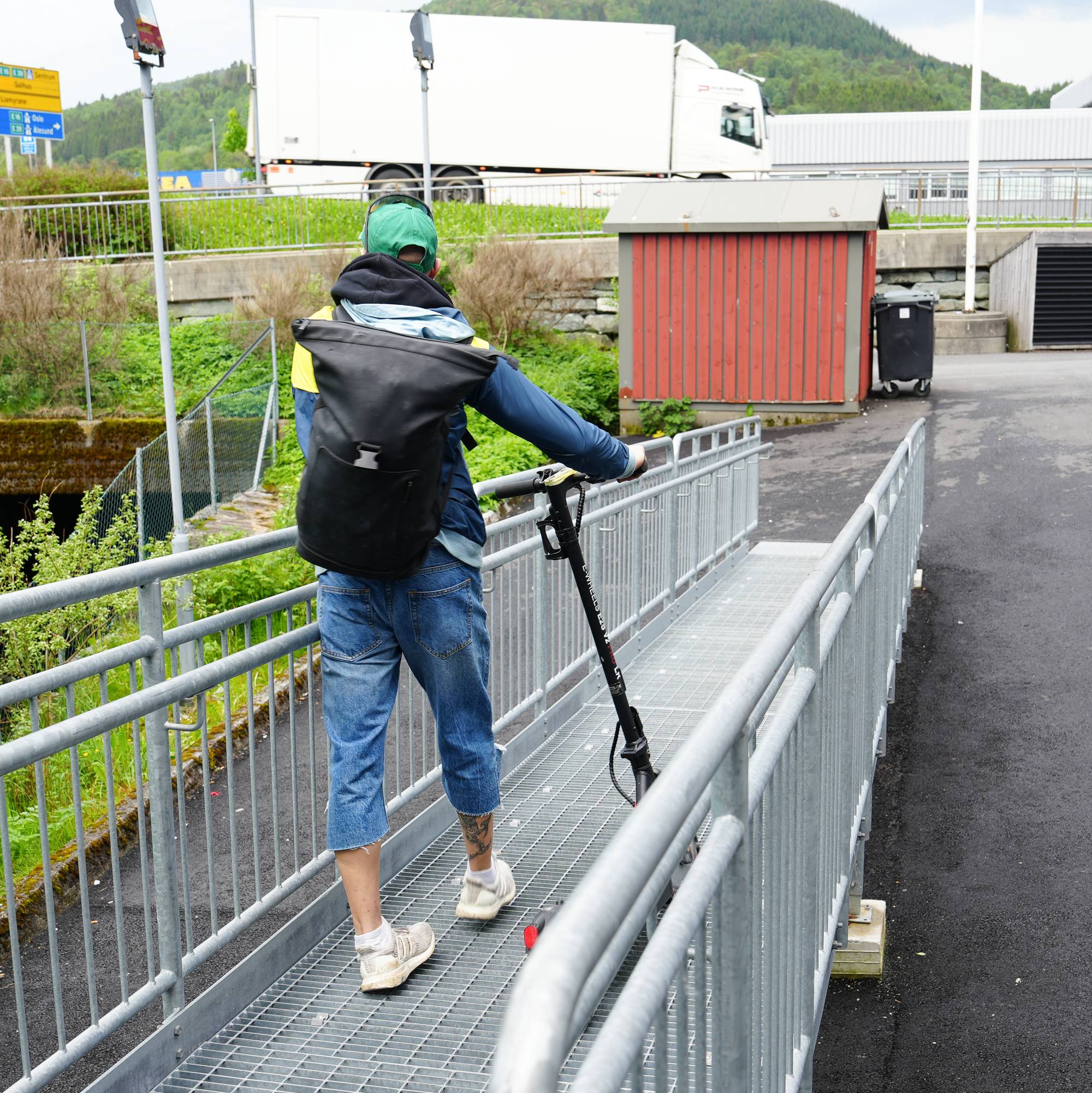 En ung mann går nedover en gangbro av stål. Han har en sort ryggsekk på ryggen, grønn caps, halvlange olabukser, hvite sokker og hvite joggesko. Han triller på en sparkesykkel. Til venstre er det grønne busker. Til høyre er det en vei, en innkjørsel. Rett fram er det et rødt skur. I bakgrunnen er det bygninger og man skimter en vei og veiskilt.