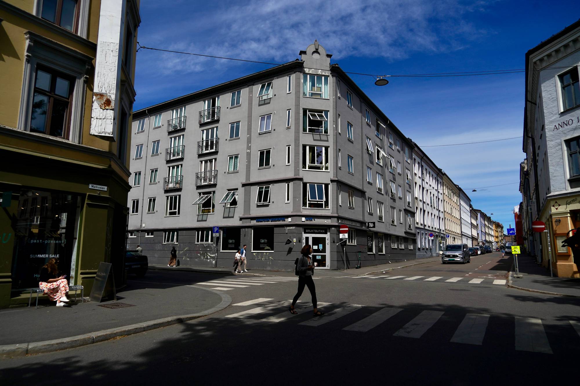En kvinne krysser et fotgjengerfelt på en gate i en by. Flere bygårder med vinduer og balkonger dominerer bildet. (Bildebeskrivelsen er laget av en KI-tjeneste)