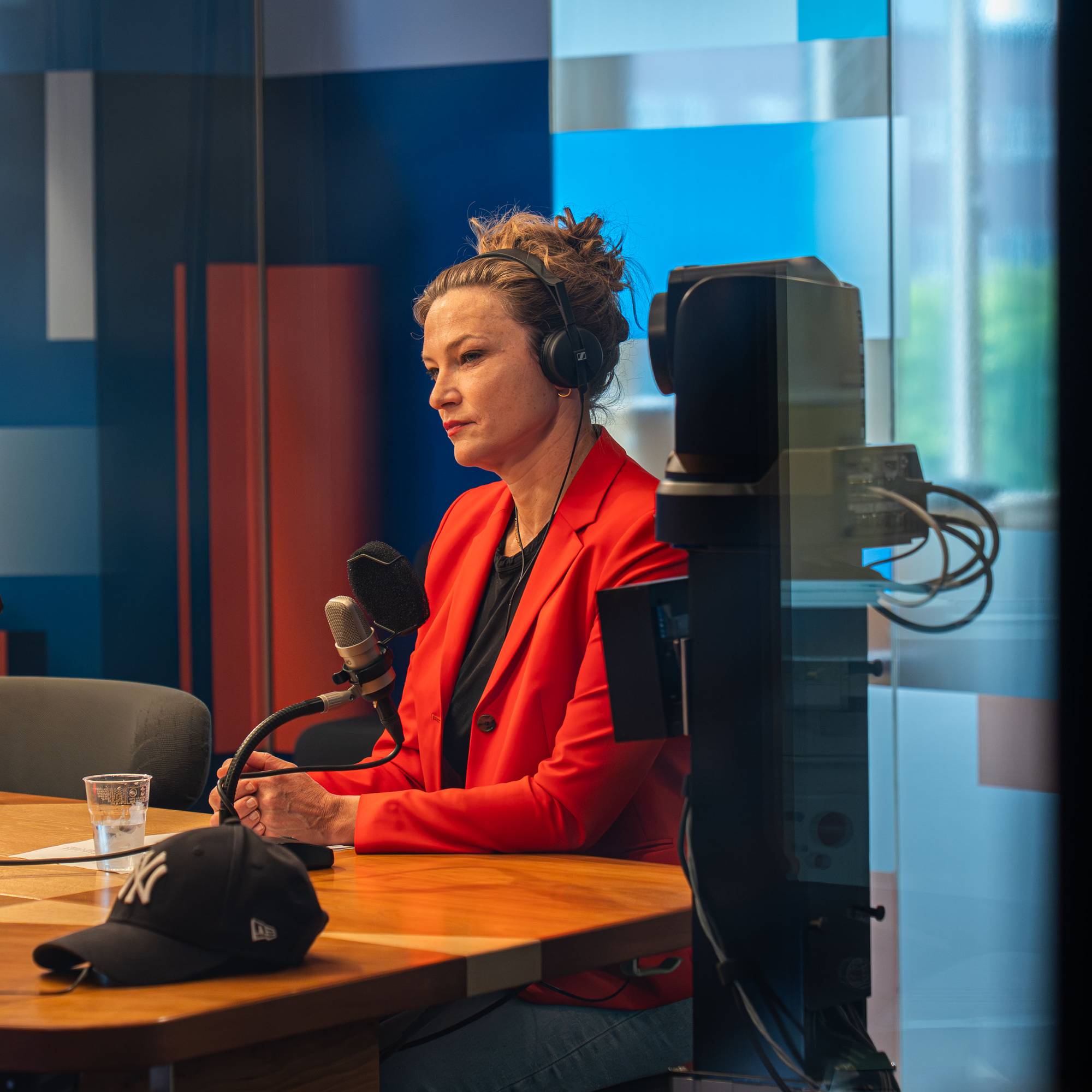 Kvinne med hodetelefoner sitter ved et bord og snakker i en mikrofon. Hun har på seg en rød blazer. I bakgrunnen er det et studio med glassvegger.