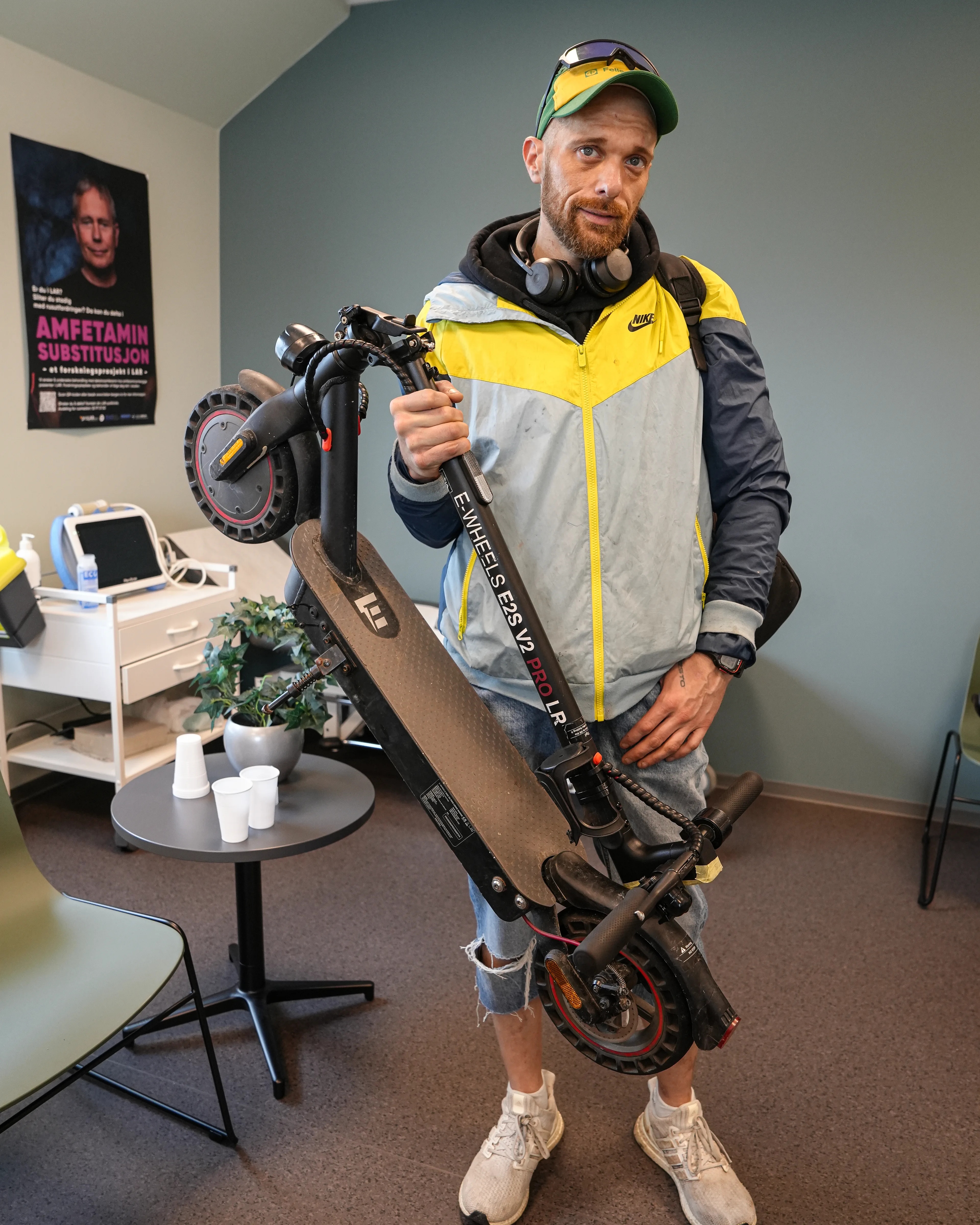 En ung mann med grønn og gul caps, grå og gul sportsjakke, halvlange olabukser og hvite joggesko står på et kontor. Han bærer en sparkesykkel i høyre hånd.