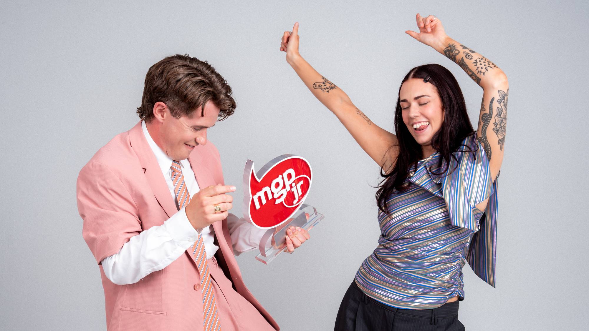 To personer ser ut til å danse. Til venstre står en mann, Martin, i rosa dress. Han holder MGPjr-trofeet. Til høyre står en kvinne, Tonje, med hendene i været. Hun har på seg en glitrende, stripete topp.