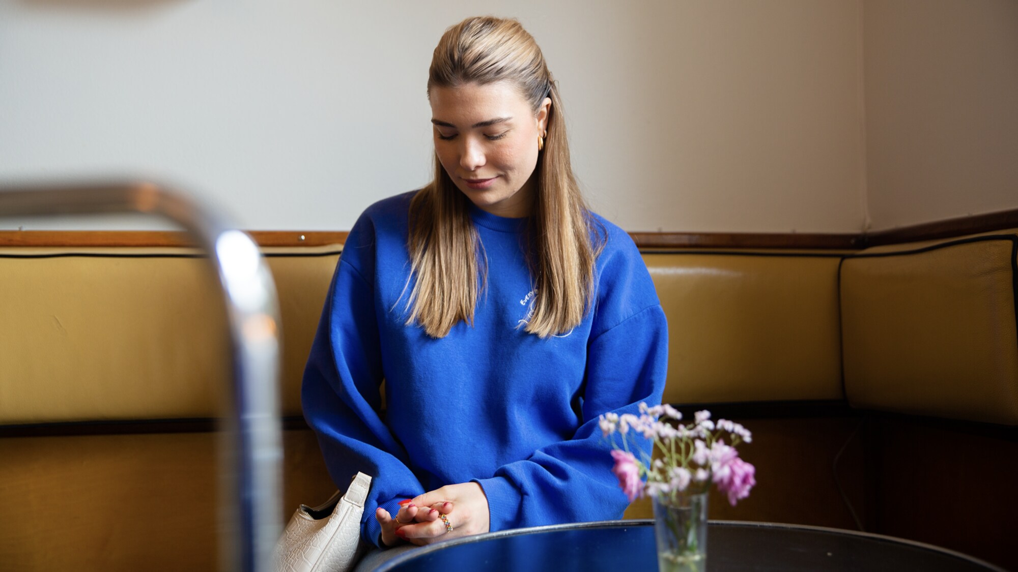 Silje sitter på en kafé og ser ned på hendene sine. Det er en tom stol på andre siden av det lille runde bordet foran henne. 