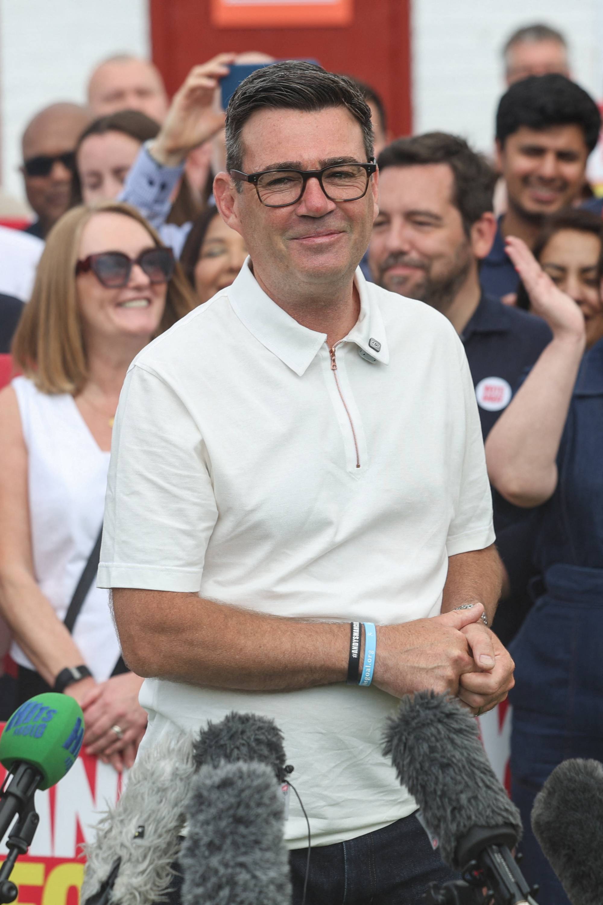 En mann med briller og hvit polo skjorte smiler mot kamera. Rundt ham står folkemengde og medier. Mannen har armene krysset og bærer to armbånd. (Bildebeskrivelsen er laget av en KI-tjeneste)