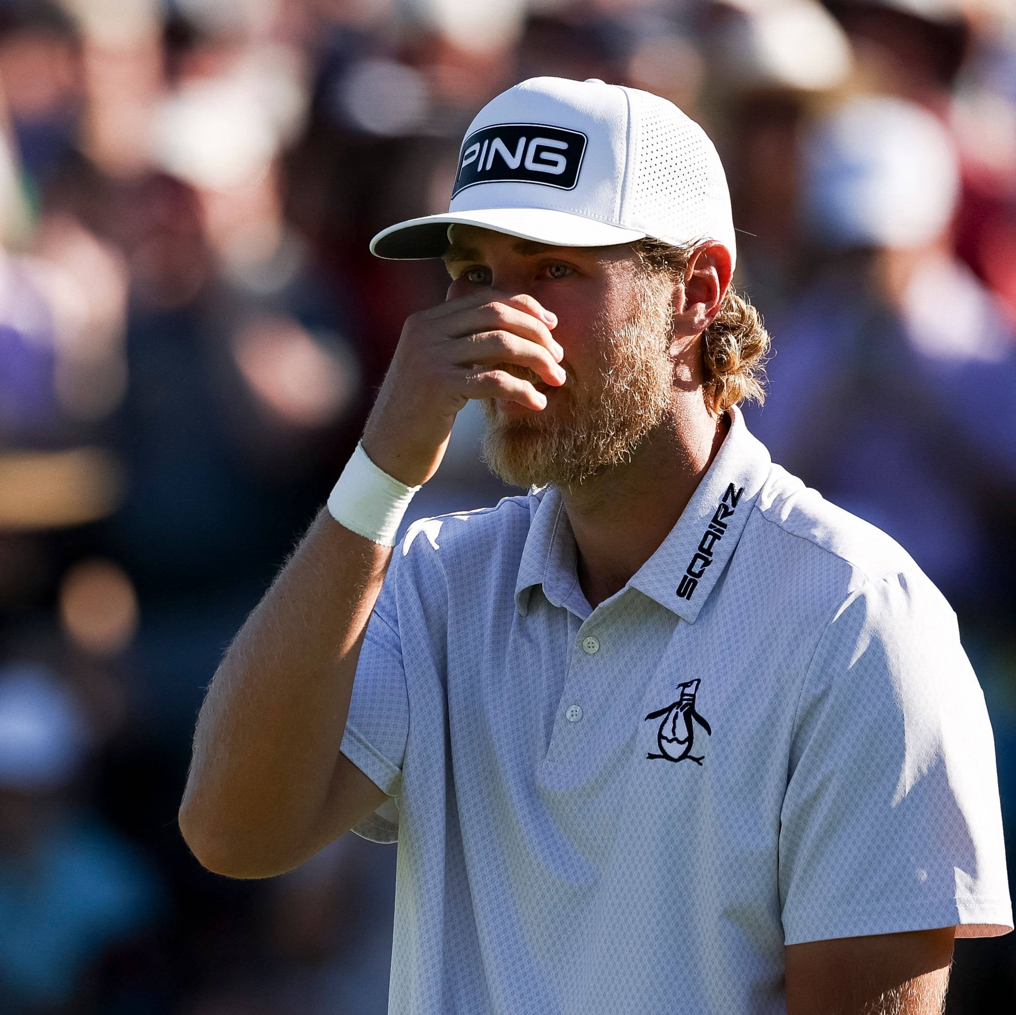 Golfer med caps og skjorte med sponsorlogoer. Mannen holder en hånd mot ansiktet. I bakgrunnen er et publikum. (Bildebeskrivelsen er laget av en KI-tjeneste)