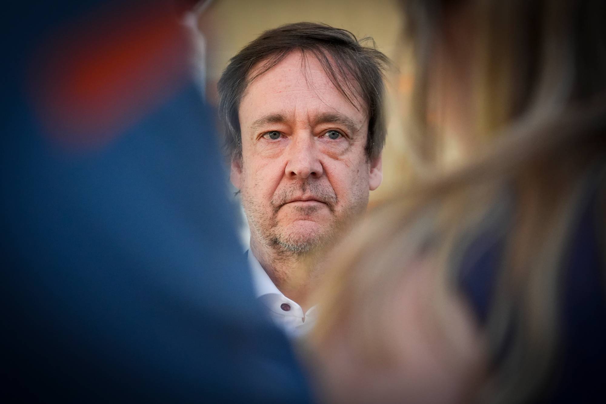 En mann i hvit skjorte ser inn i kamera. Han er omringet av uskarpe personer. Blå og oransje farger dominerer forgrunnen. Bildet er tatt utendørs. (Bildebeskrivelsen er laget av en KI-tjeneste)