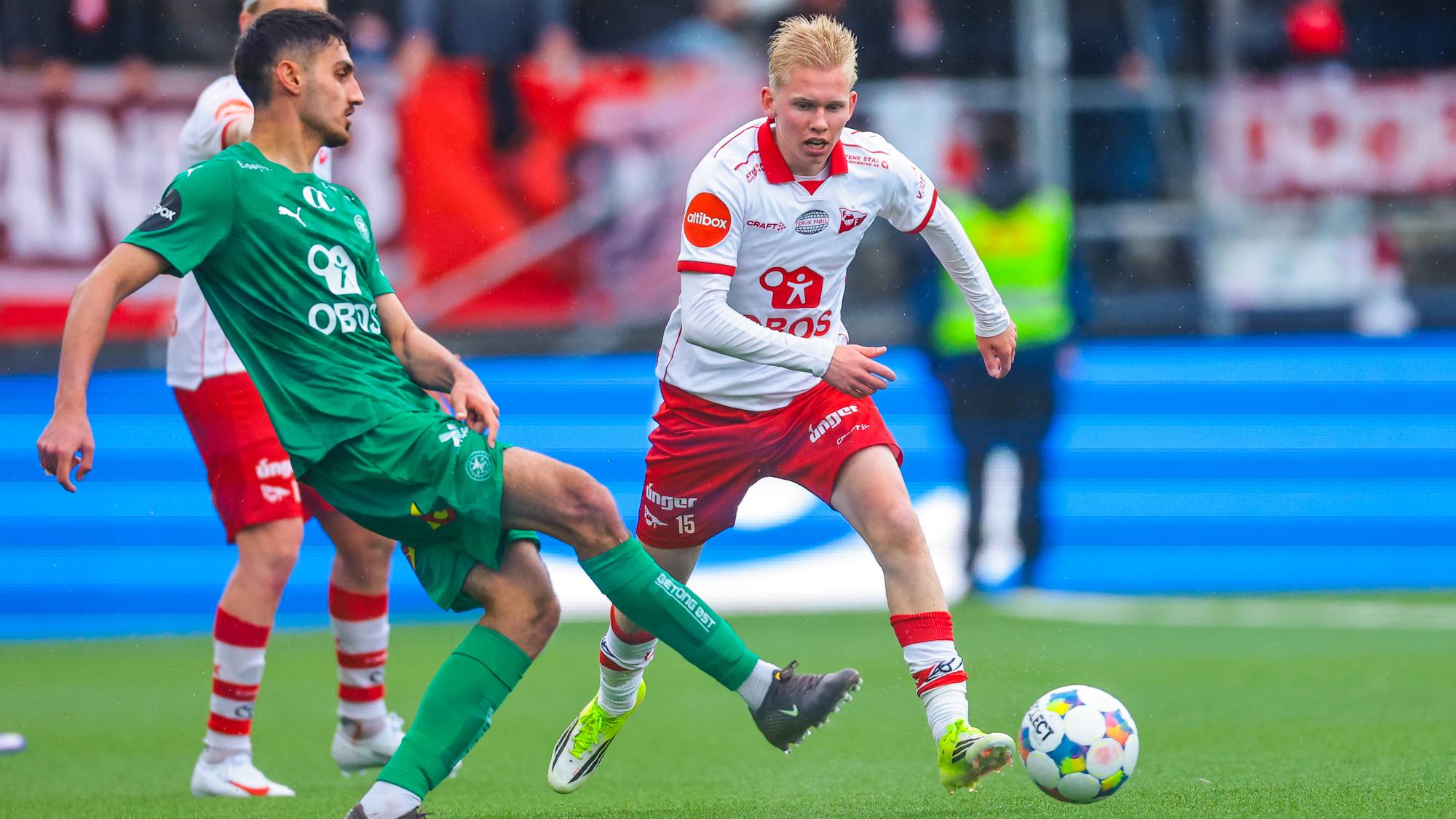 To fotballspillere i kamp på et grønt gressfelt. Den ene spilleren, iført grønn drakt, prøver å takle ballen fra den andre spilleren. Spilleren i grønt har mørkt hår. Spilleren i hvitt og rødt har blondt hår. Kampen foregår på en arena med tilskuere i bakgrunnen. (Bildebeskrivelsen er laget av en KI-tjeneste)