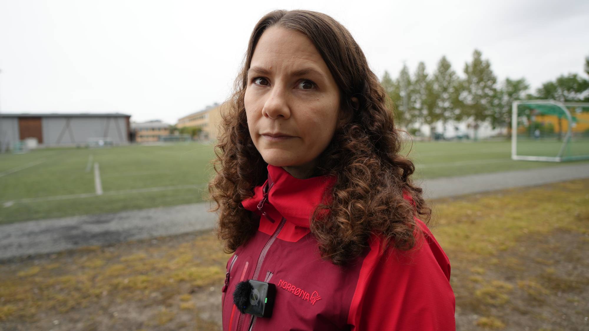 Kvinne med krøllete hår og rød jakke står på en fotballbane. (Bildebeskrivelsen er laget av en KI-tjeneste)