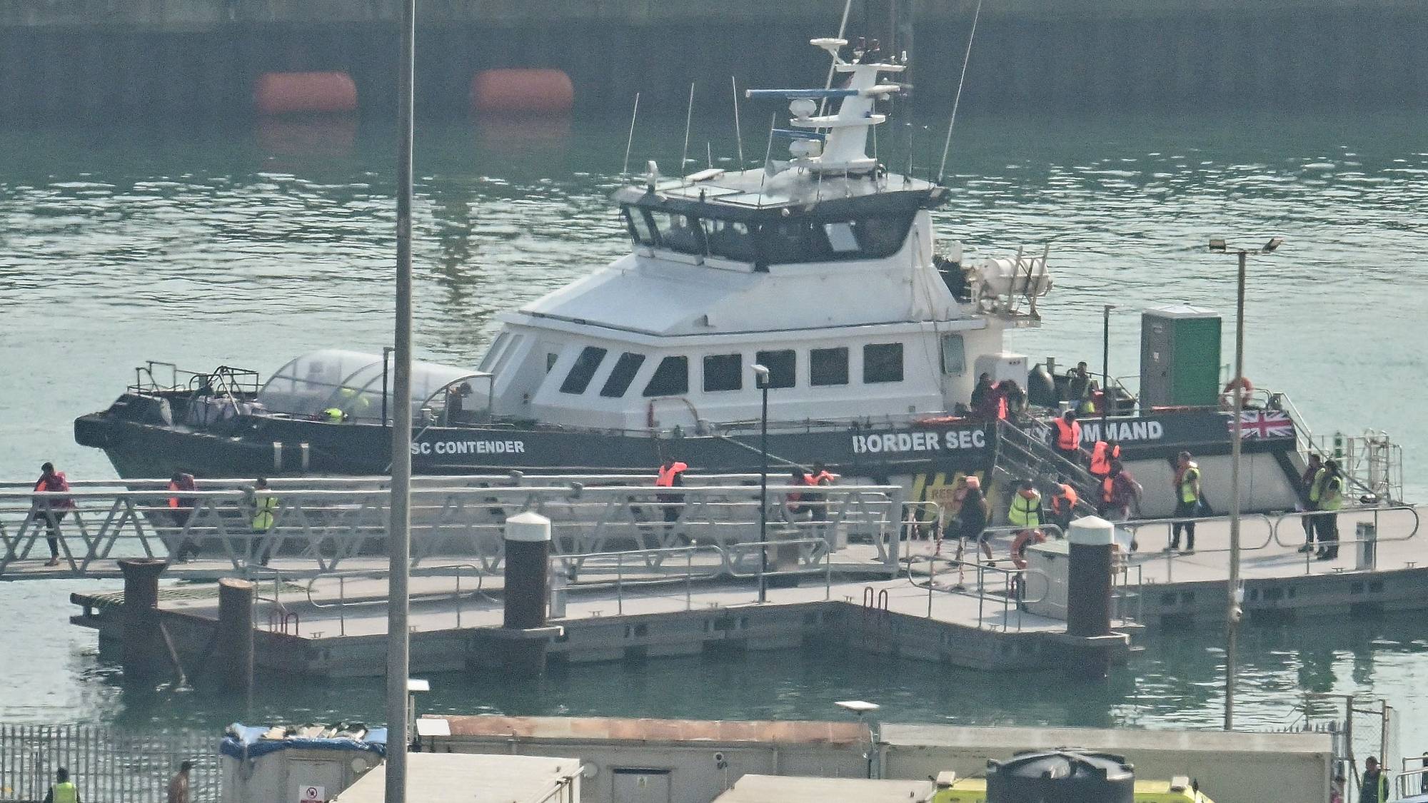Personer med redningsvester går i land fra et grensekontrollfartøy til en brygge. (Bildebeskrivelsen er laget av en KI-tjeneste)