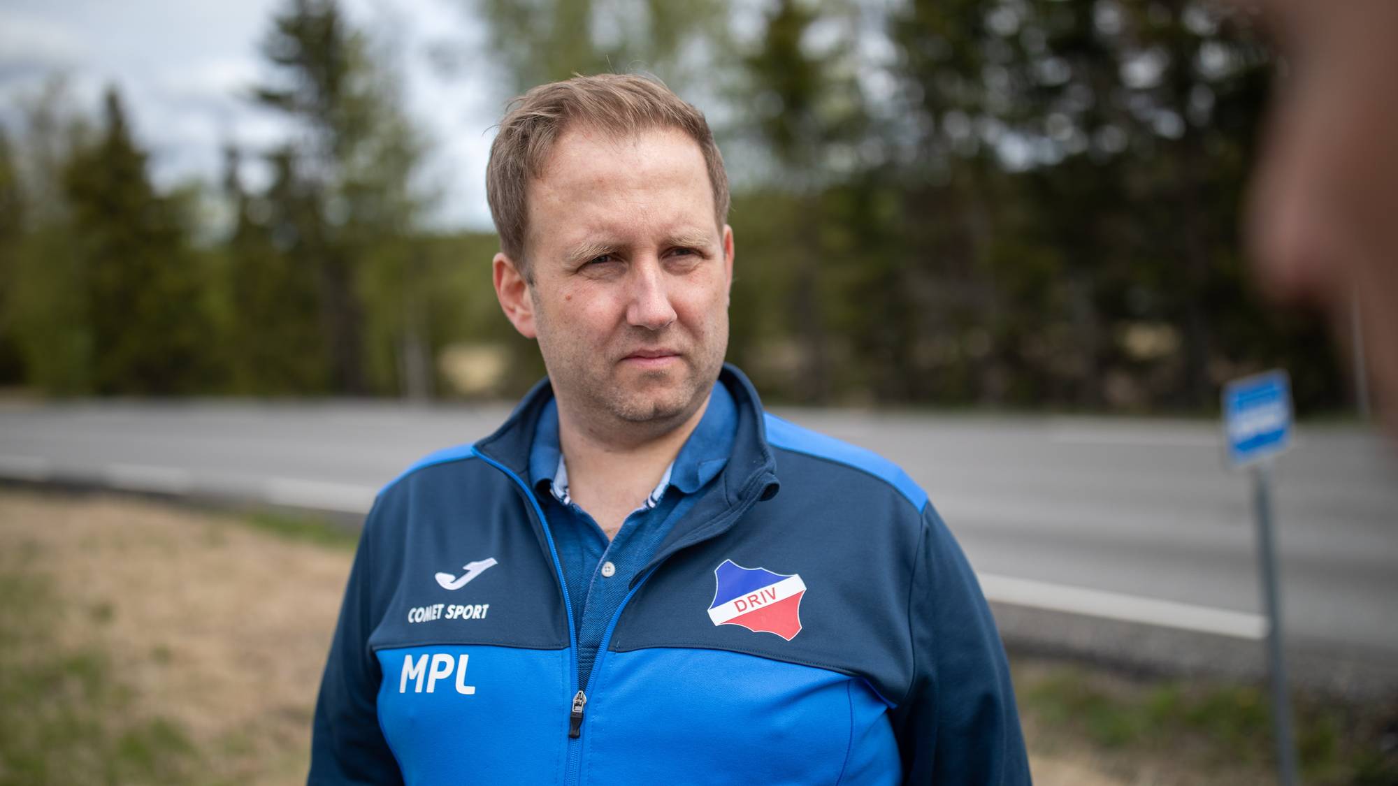 En mann i blå jakke står utendørs. Jakken har logoer og tekst. I bakgrunnen er det trær og en vei. (Bildebeskrivelsen er laget av en KI-tjeneste)
