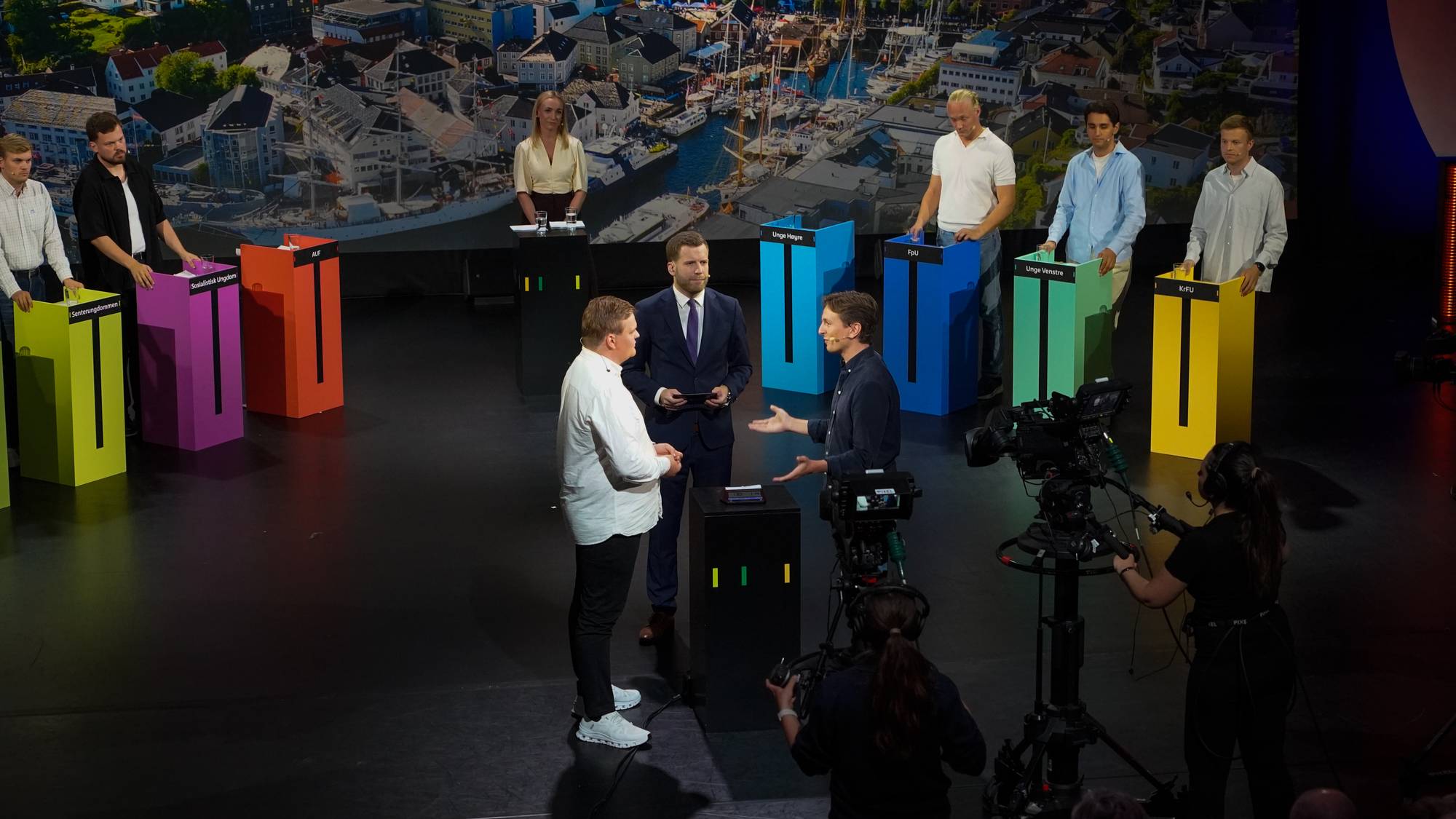 Skjervø og Svenneby debatterer midt i bildet. Rundt dem står de andre ungdomspolitikerne og følger med. Settingen er en typisk debattsituasjon, der bildet viser de forskjellige debattantene som står bak hver sin talerstol, i det som skal representere partienes farge.