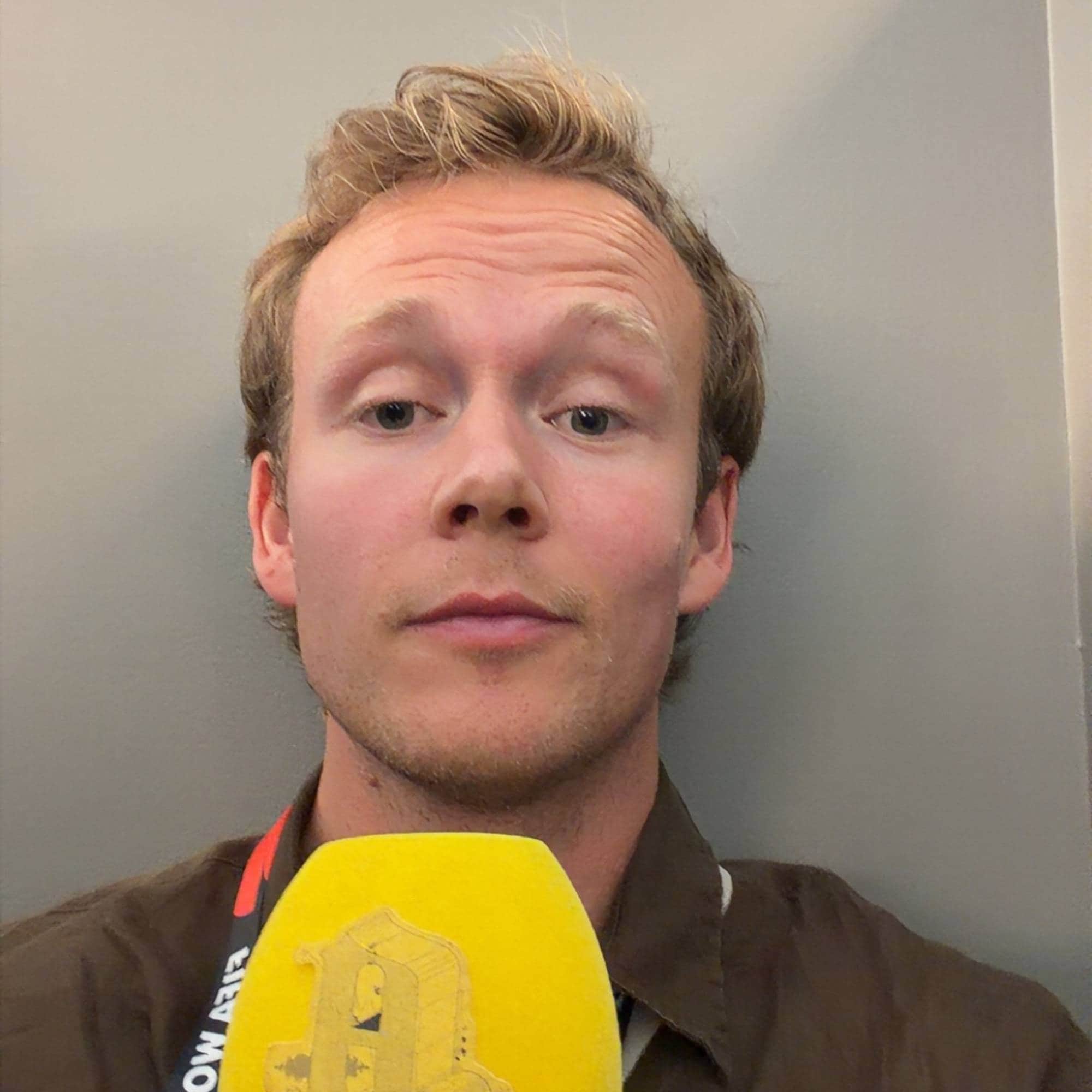 Mann ser i kamera. Holder en mikrofon foran seg med en gul aftenposten hette på mikrofonen.