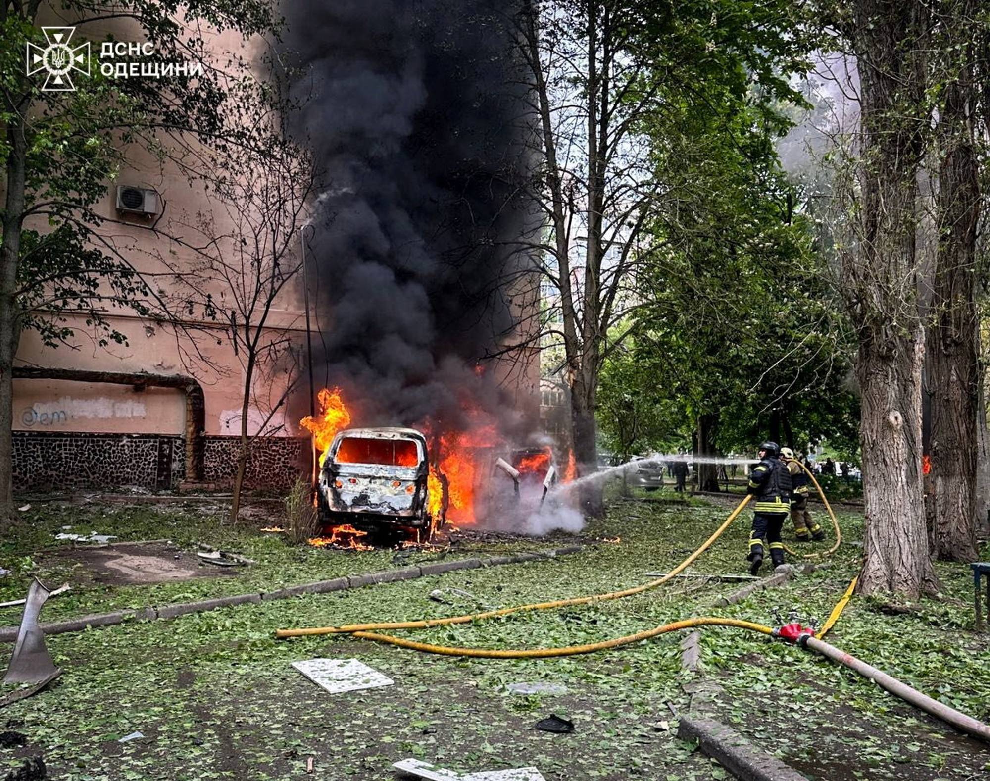 Brannmannskaper slukker en bil som brenner kraftig. Bilen står parkert i et grønt område med trær og vegetasjon. En bygning er synlig i bakgrunnen. Flere brannslanger ligger på bakken, og det er mye røyk i luften. (Bildebeskrivelsen er laget av en KI-tjeneste)
