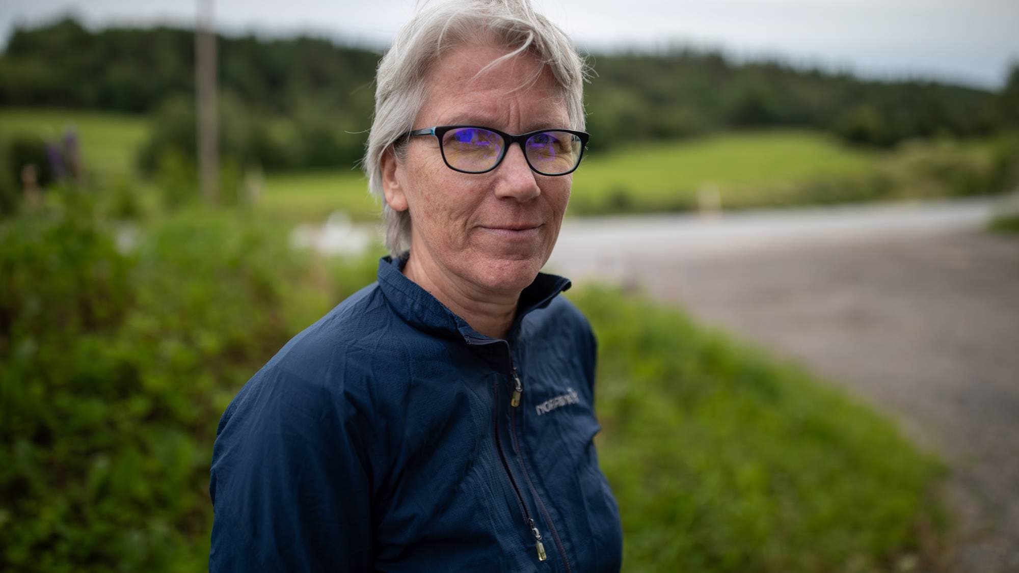 kvinne med kort grått hår, og sorte briller, 
Hun er vendt mot venstre men ser rett inn i kamera.
