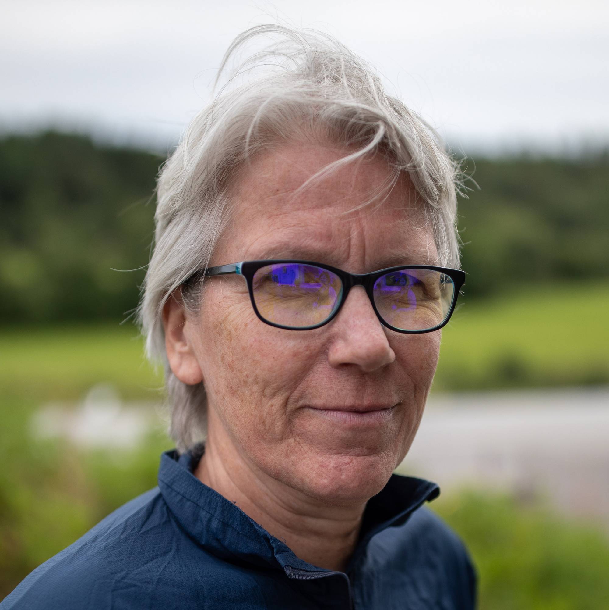 kvinne med kort grått hår, og sorte briller, 
Hun er vendt mot venstre men ser rett inn i kamera.