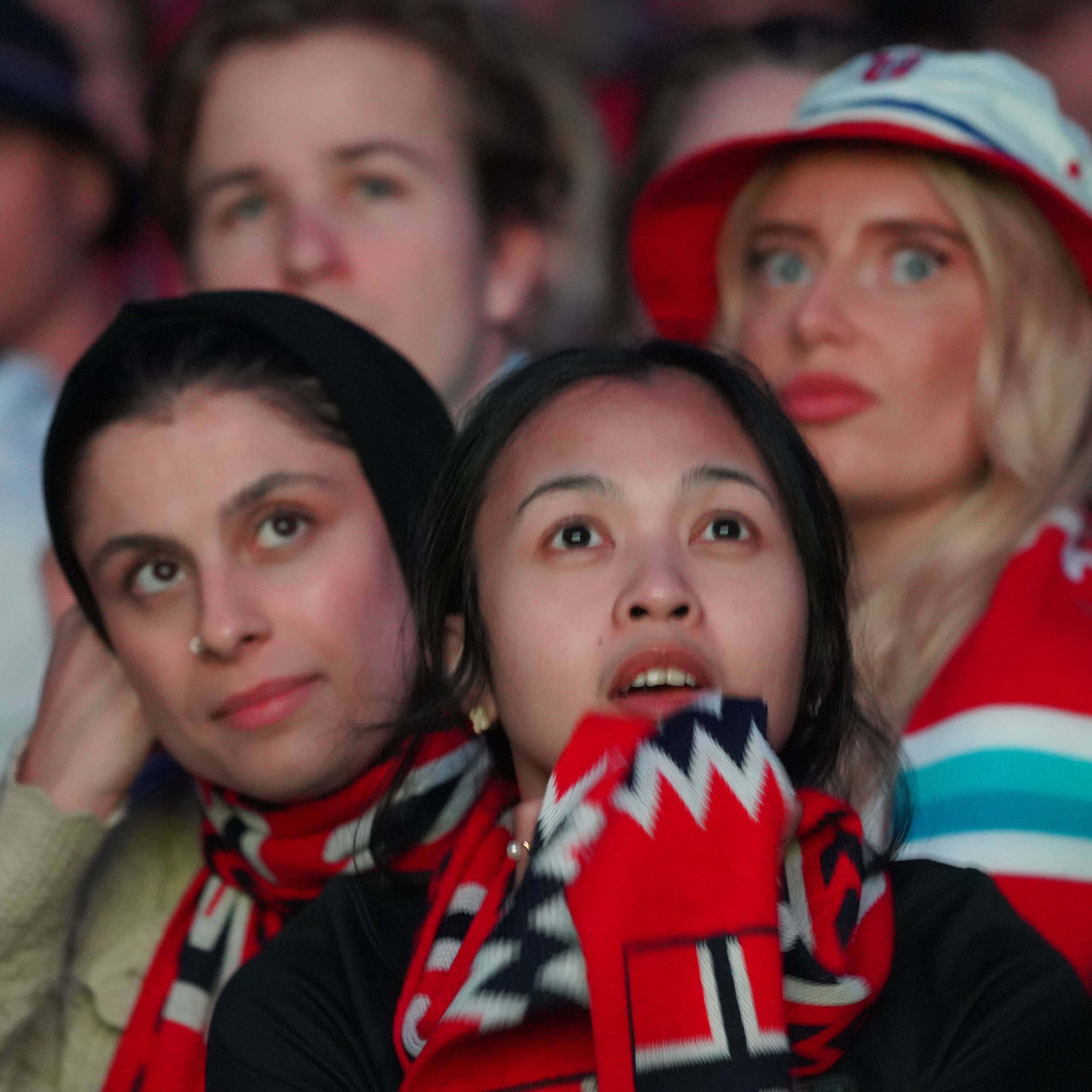 Flere personer ser opp i et bilde. En kvinne med mørkt hår og skjerf har hånden ved tinningen. En annen kvinne med langt, lyst hår har på seg en rød og hvit hatt. De sitter på en tribuna og har på seg supporterutstyr.