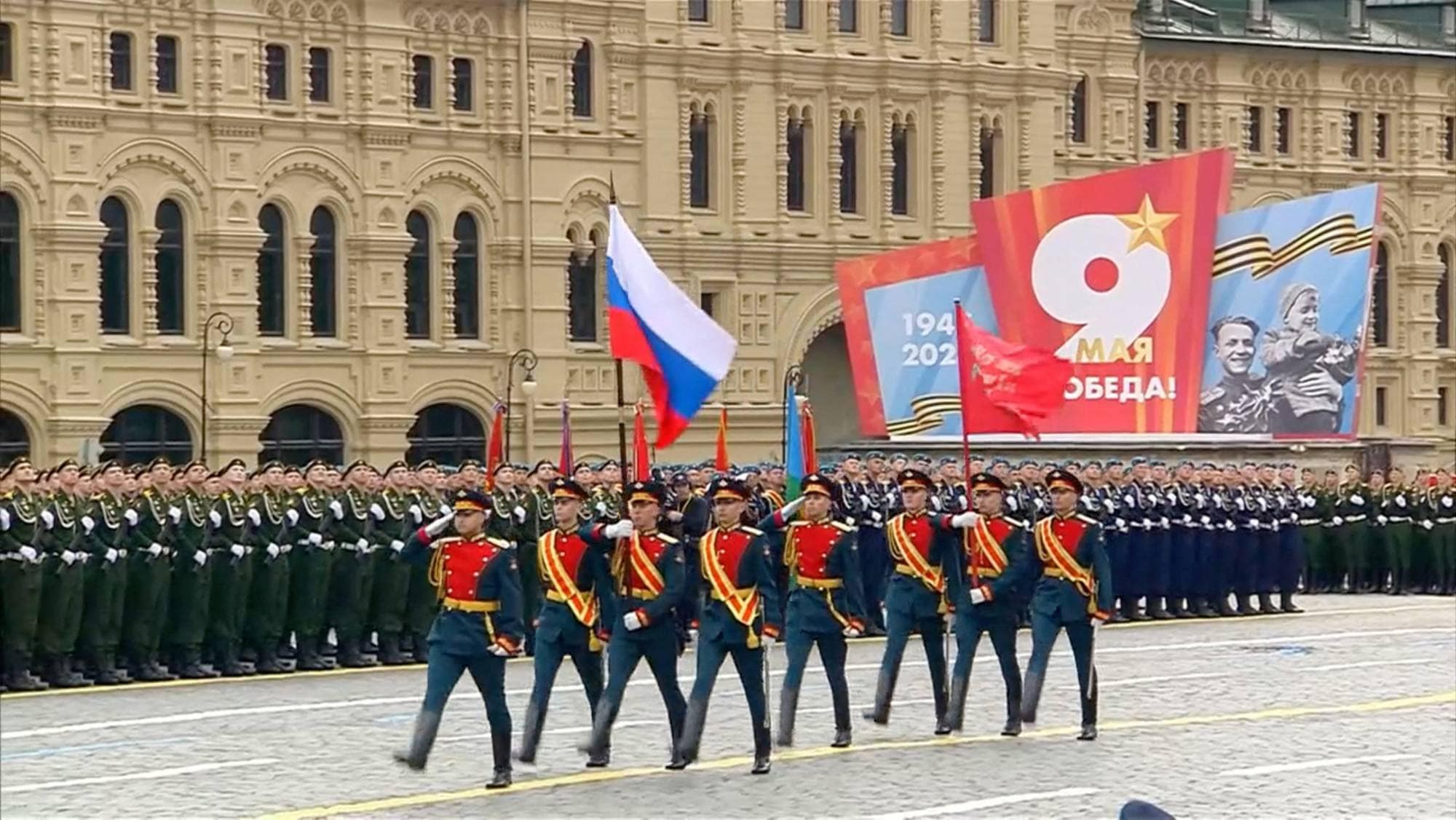 Militærparade på Den røde plass i Moskva. Soldater i paradeuniformer marsjerer forbi med russiske flagg. Bak soldatene står rekker av soldater i grønne uniformer. I bakgrunnen er det store bannere med russiske inskripsjoner og bilder, og en detaljert bygning. (Bildebeskrivelsen er laget av en KI-tjeneste)