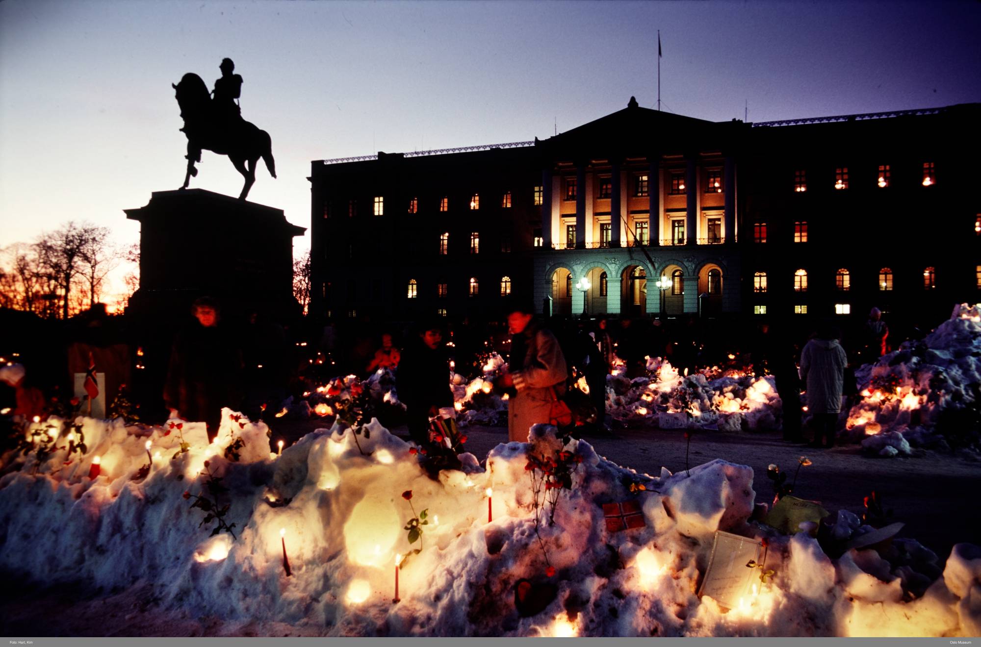 En snødekket Slottsplassen er fylt med blomster og lys, og sørgende går forbi. I forgrunnen sees rytterstatuen, og i bakgrunnen er Slottet i kveldslys.
