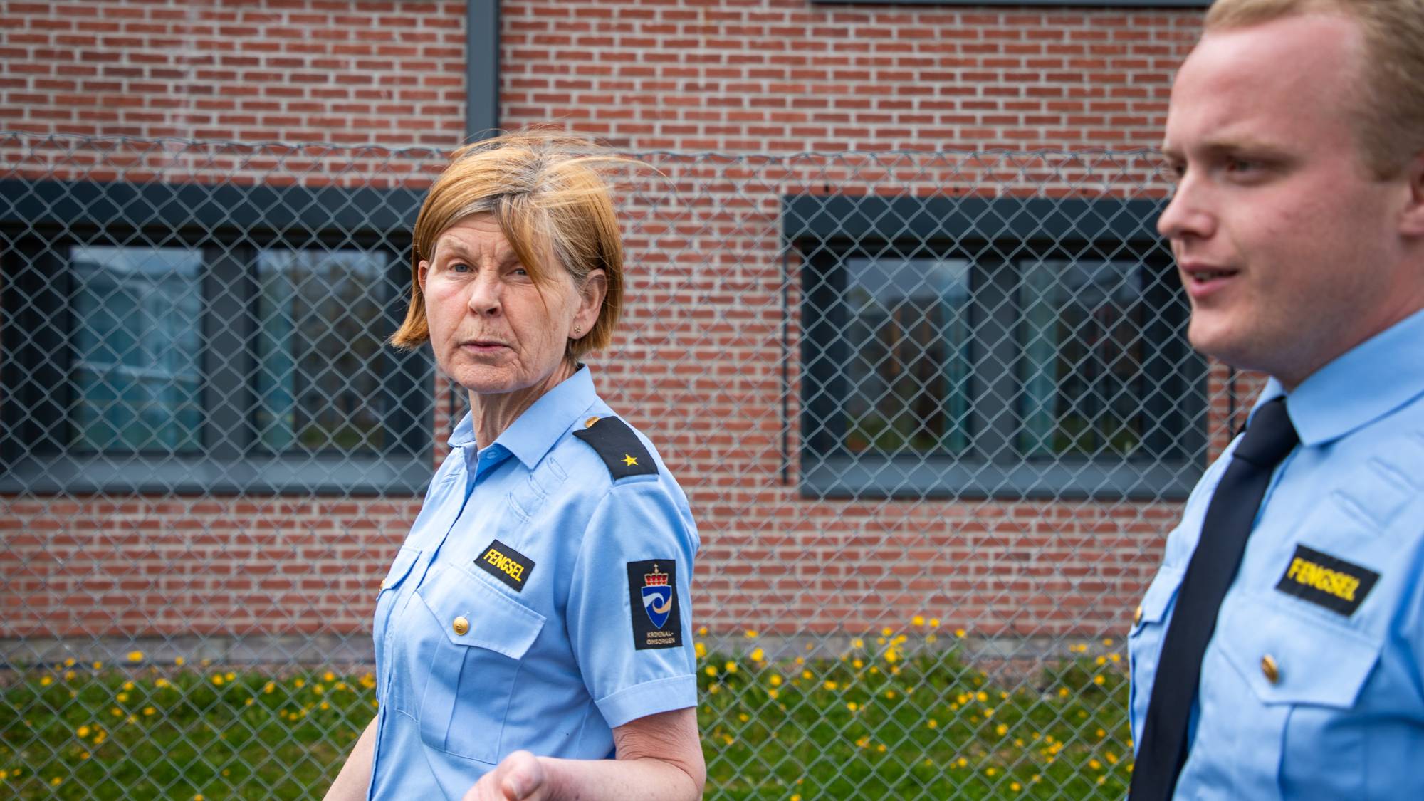 Bildet viser to fengselsbetjenter som går forbi et murbygg bak et gittergjerde