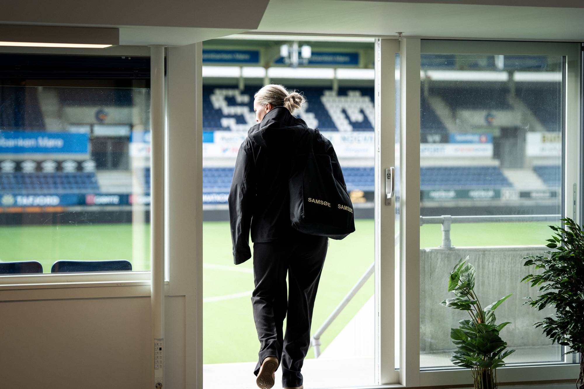 En kvinne i svart jakke med veske går ut på en fotballstadion sett fra innsiden av et rom. (Bildebeskrivelsen er laget av en KI-tjeneste)
