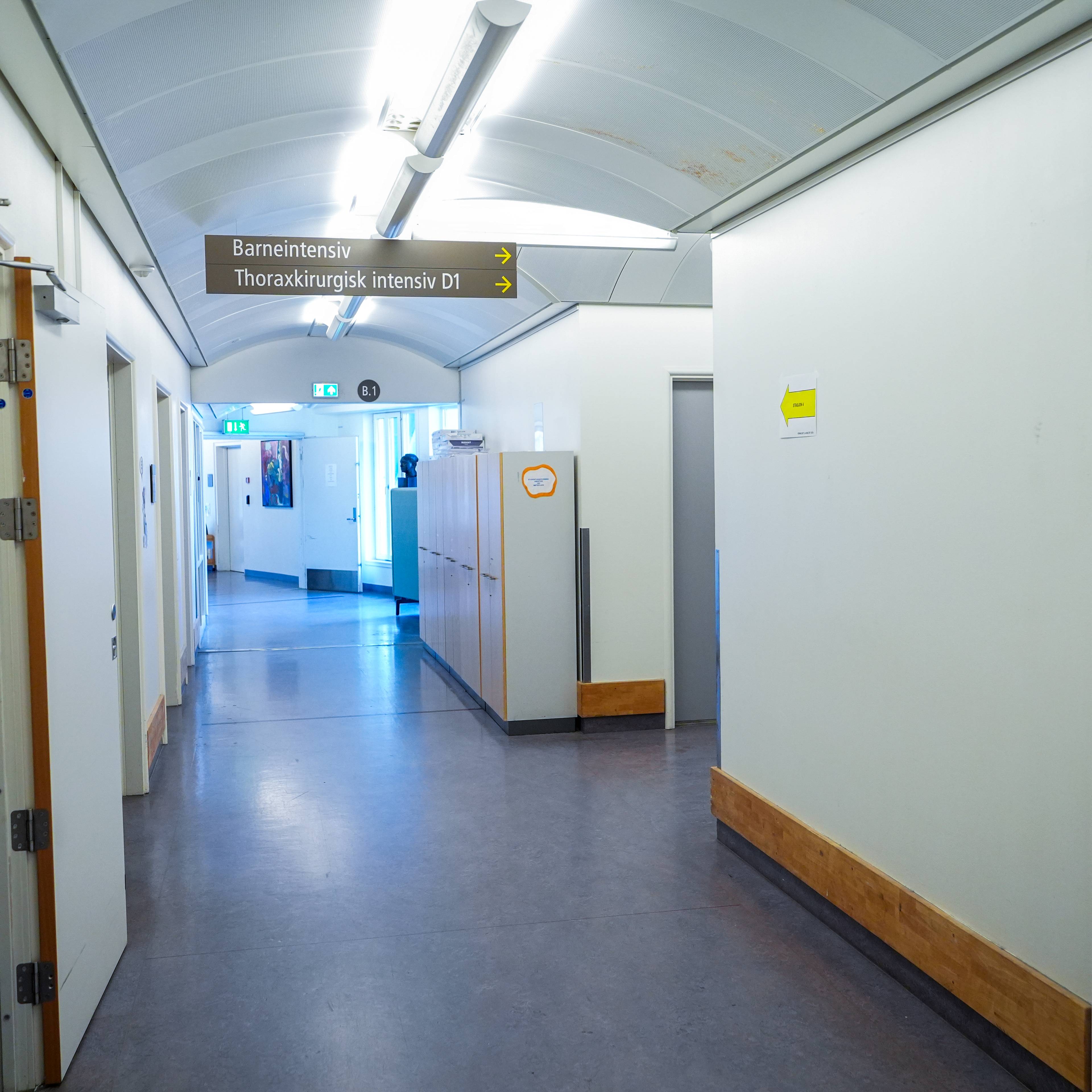 Sykehuskorridor med skilt for Barneintensiv og Thoraxkirurgisk intensiv. Korridoren er opplyst av lysstoffrør. På venstre side av korridoren står en skriver. (Bildebeskrivelsen er laget av en KI-tjeneste)