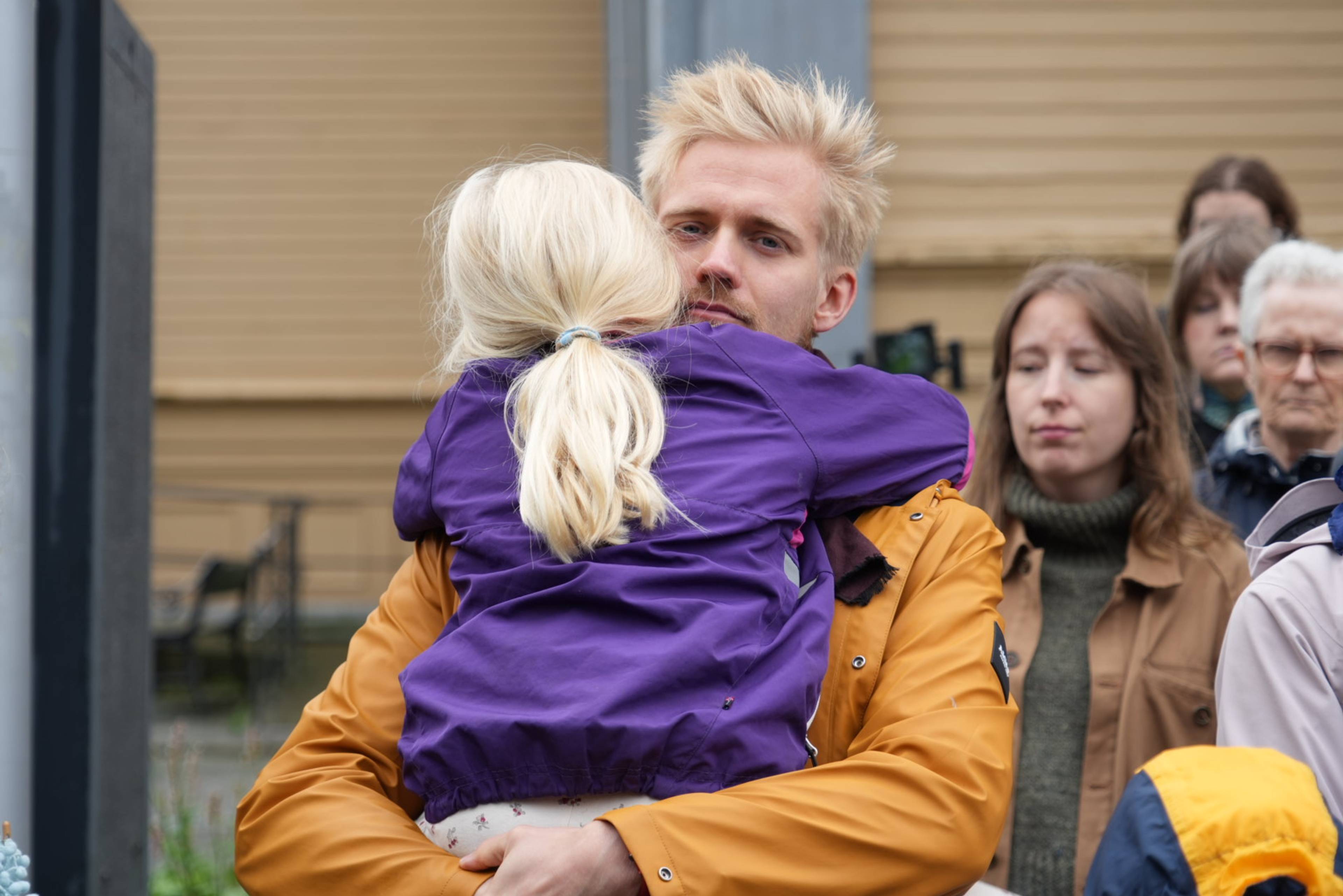 En mann i gul jakke holder et barn i lilla jakke i en omfavnelse, med folk i bakgrunnen.