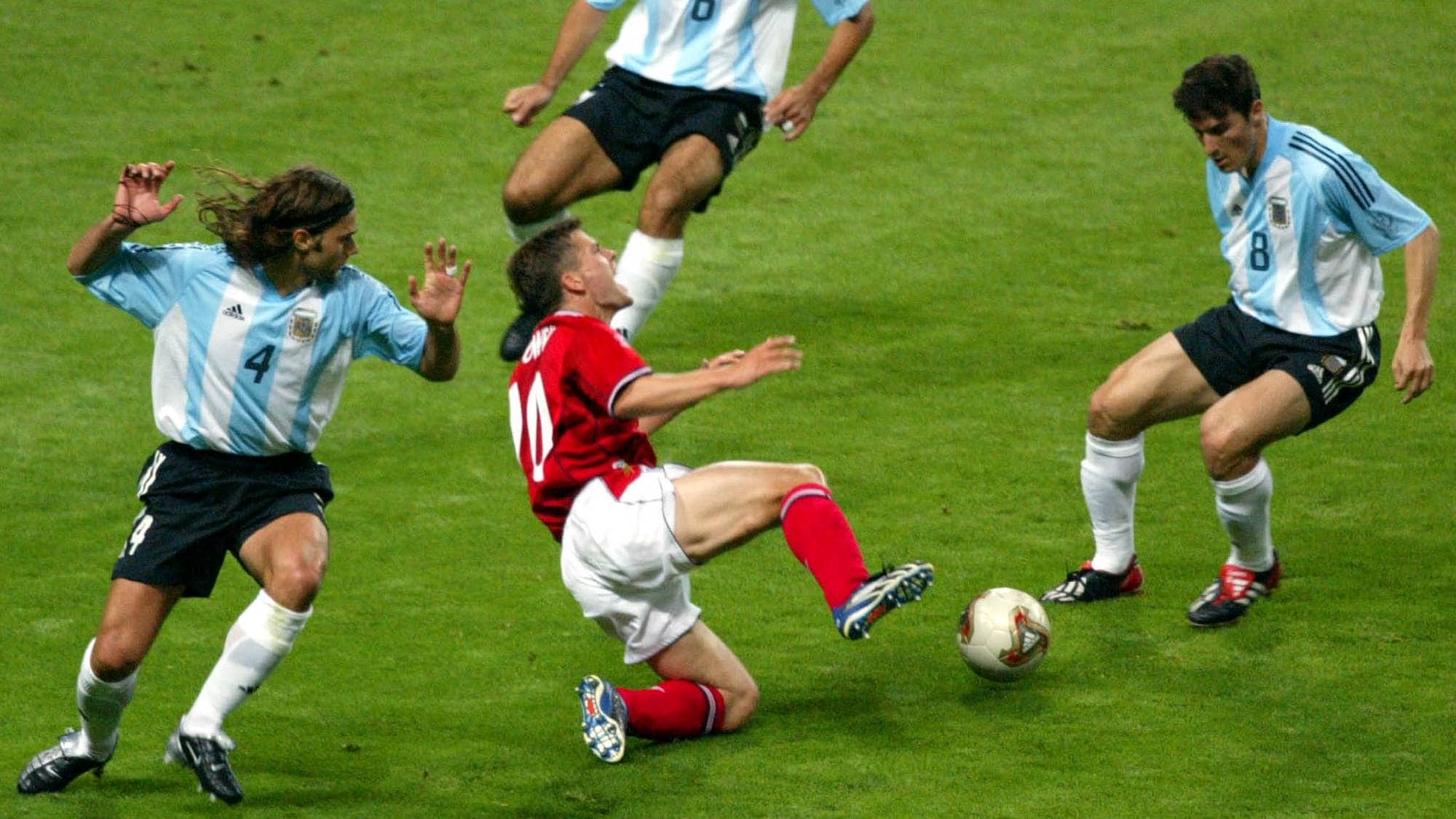 Tre fotballspillere fra Argentina omringer en spiller i rød drakt på en gressbane. (Bildebeskrivelsen er laget av en KI-tjeneste)