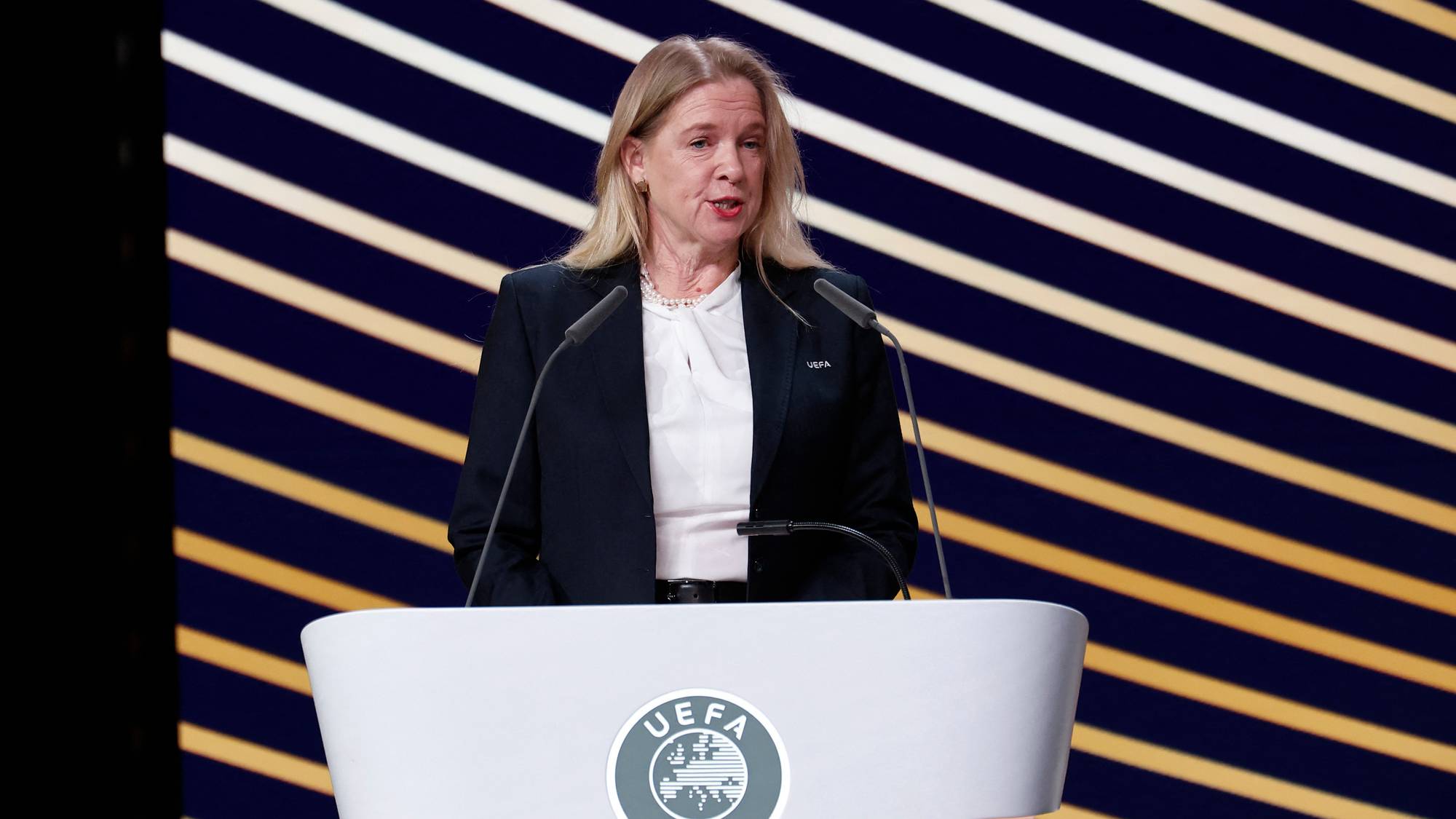 Kvinne i mørk dress taler fra en talerstol med UEFA-logo. Bakgrunnen har gule og mørke striper. (Bildebeskrivelsen er laget av en KI-tjeneste)