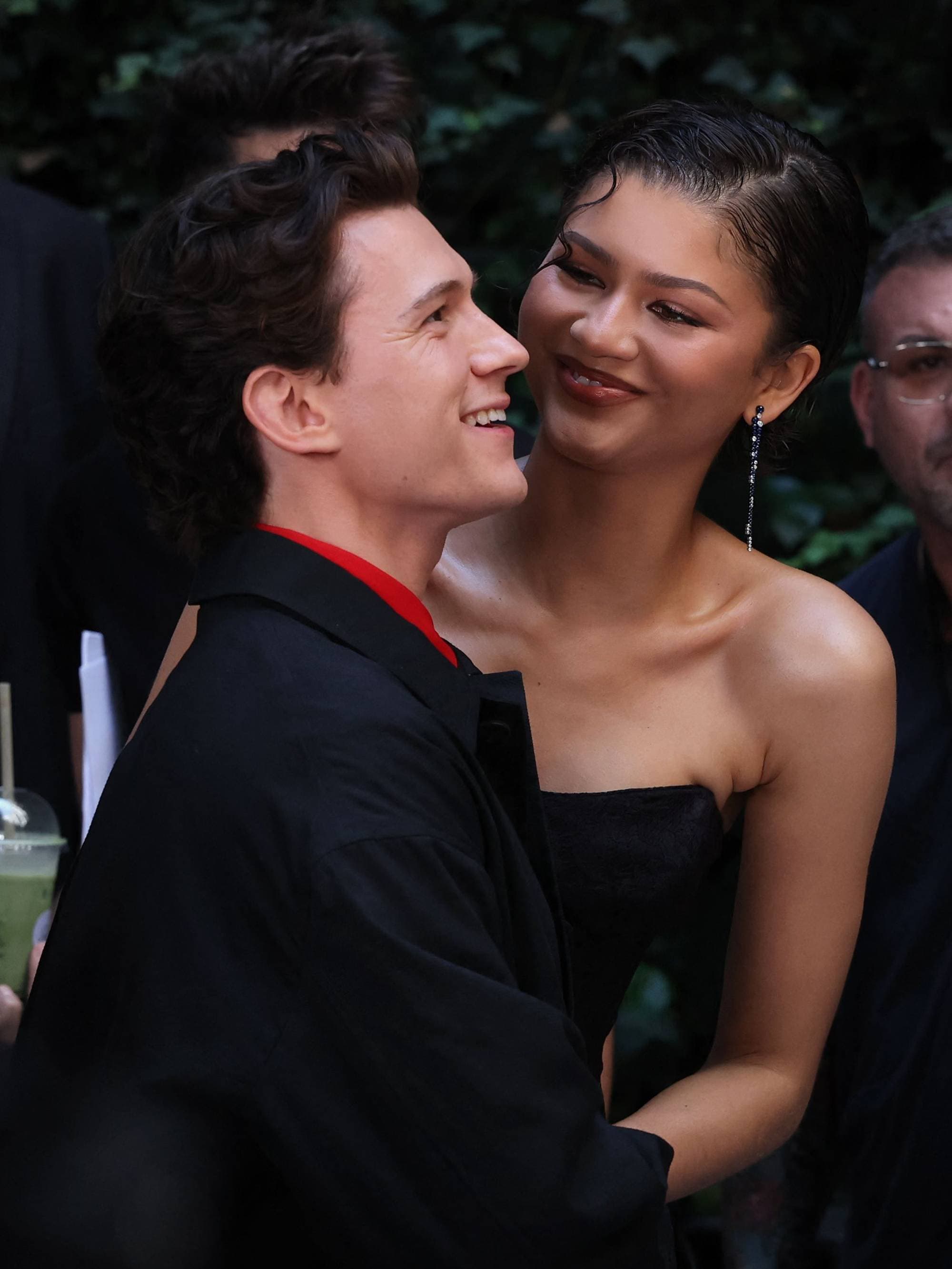 Skuespillerne Tom Holland og Zendaya smiler til hverandre. De er i en utendørs setting med andre personer synlig i bakgrunnen. Holland er ikledd en svart jakke over en rød skjorte, mens Zendaya bærer en stroppeløs svart kjole. (Bildebeskrivelsen er laget av en KI-tjeneste)