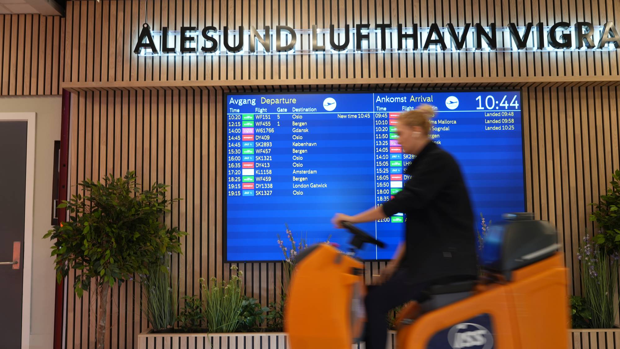 Informasjonstavle med flyavganger på Ålesund lufthavn Vigra med en person på vaskemaskin i forgrunnen. (Bildebeskrivelsen er laget av en KI-tjeneste)