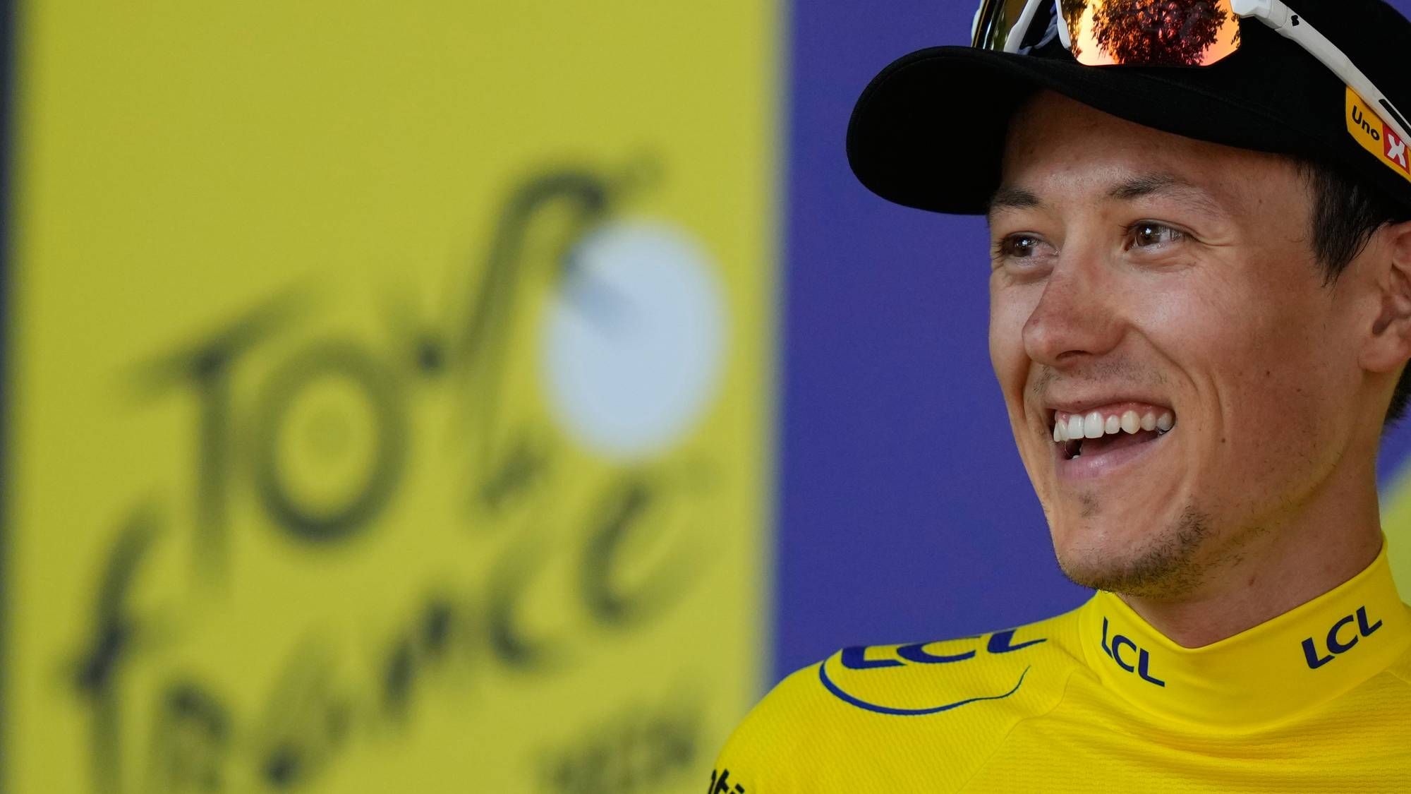 Syklist i gul ledertrøye fra Tour de France smiler foran en gul bakgrunn med rittets logo. (Bildebeskrivelsen er laget av en KI-tjeneste)