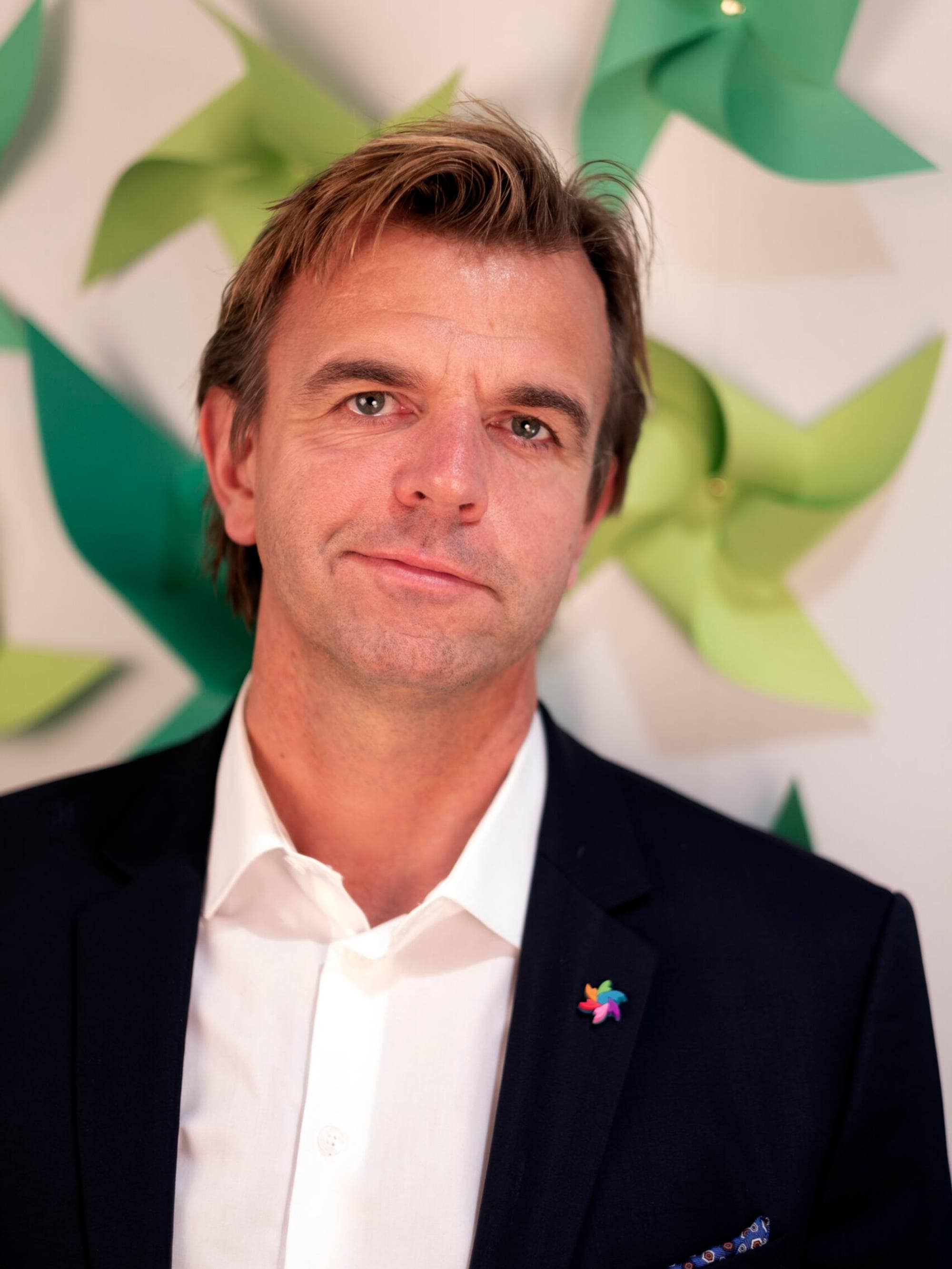 Portrett av mann i mørk dress foran en vegg dekorert med grønne papirvindmøller. (Bildebeskrivelsen er laget av en KI-tjeneste)