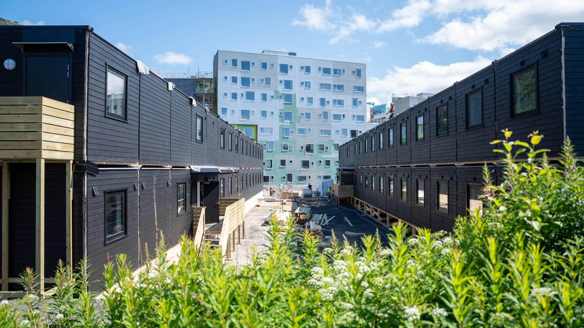Brakkerigg i svart treverk står på kvar side av ein byggeplass med ei større boligblokk i bakgrunnen.